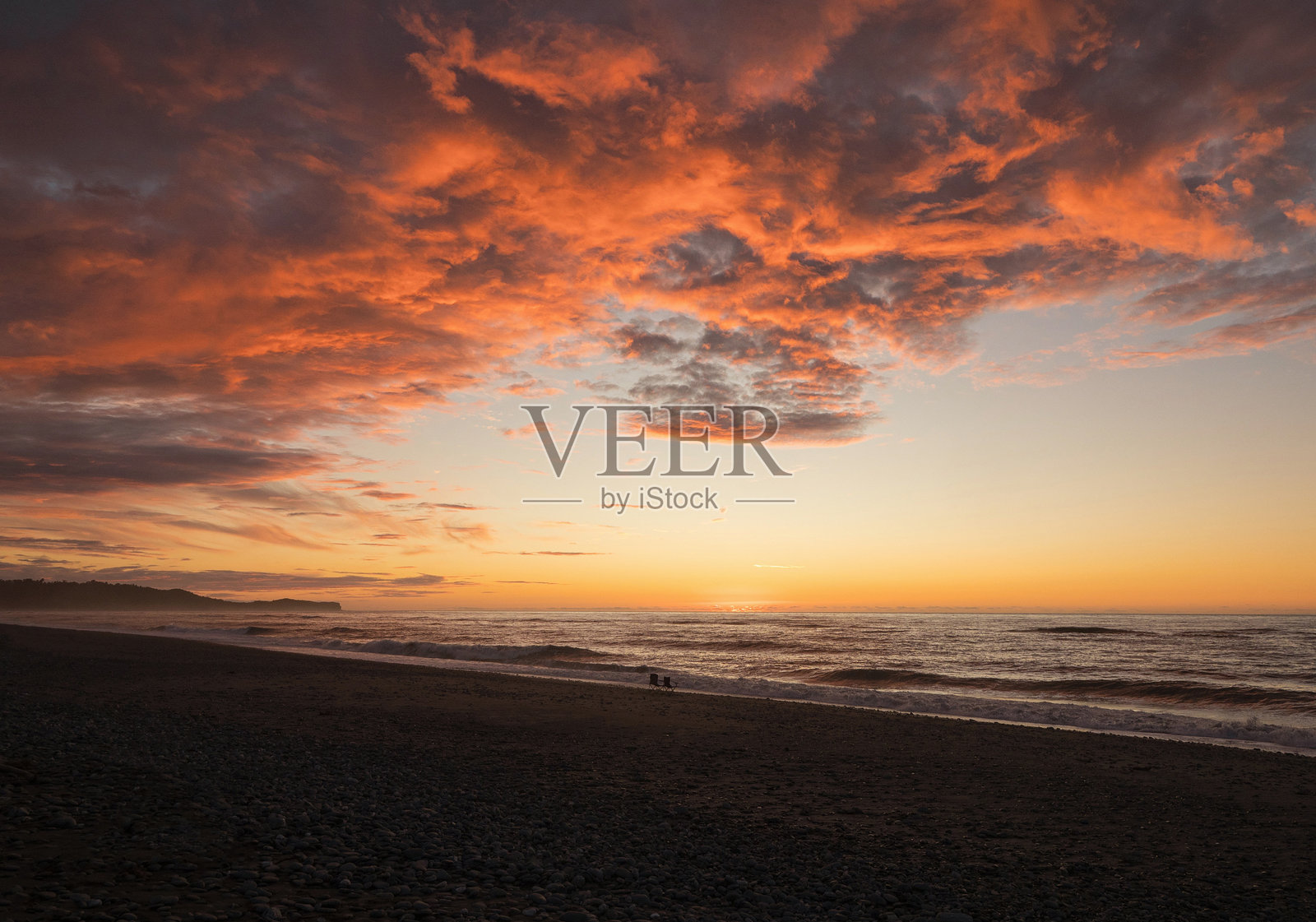 新西兰南岛西海岸吉莱斯比海滩上，两把空的露营椅，背景是海浪和五彩斑斓的晚霞。照片摄影图片
