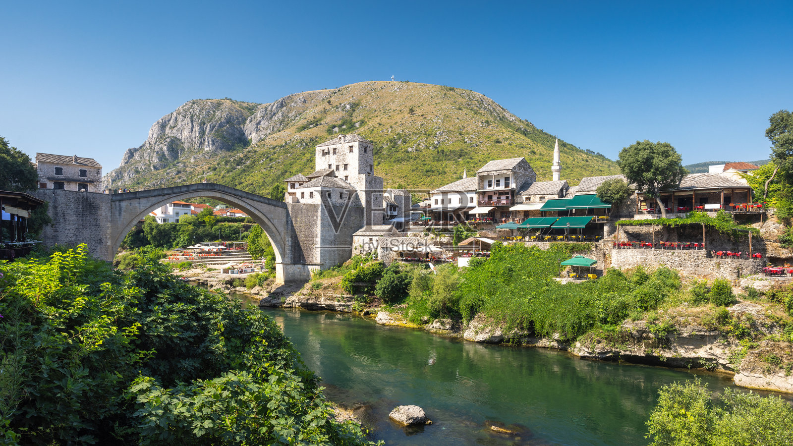 波斯尼亚和黑塞哥维那莫斯塔尔镇的斯塔里桥，横跨宁静的内雷特瓦河，周围有历史建筑、郁郁葱葱的绿树和山脉背景。照片摄影图片