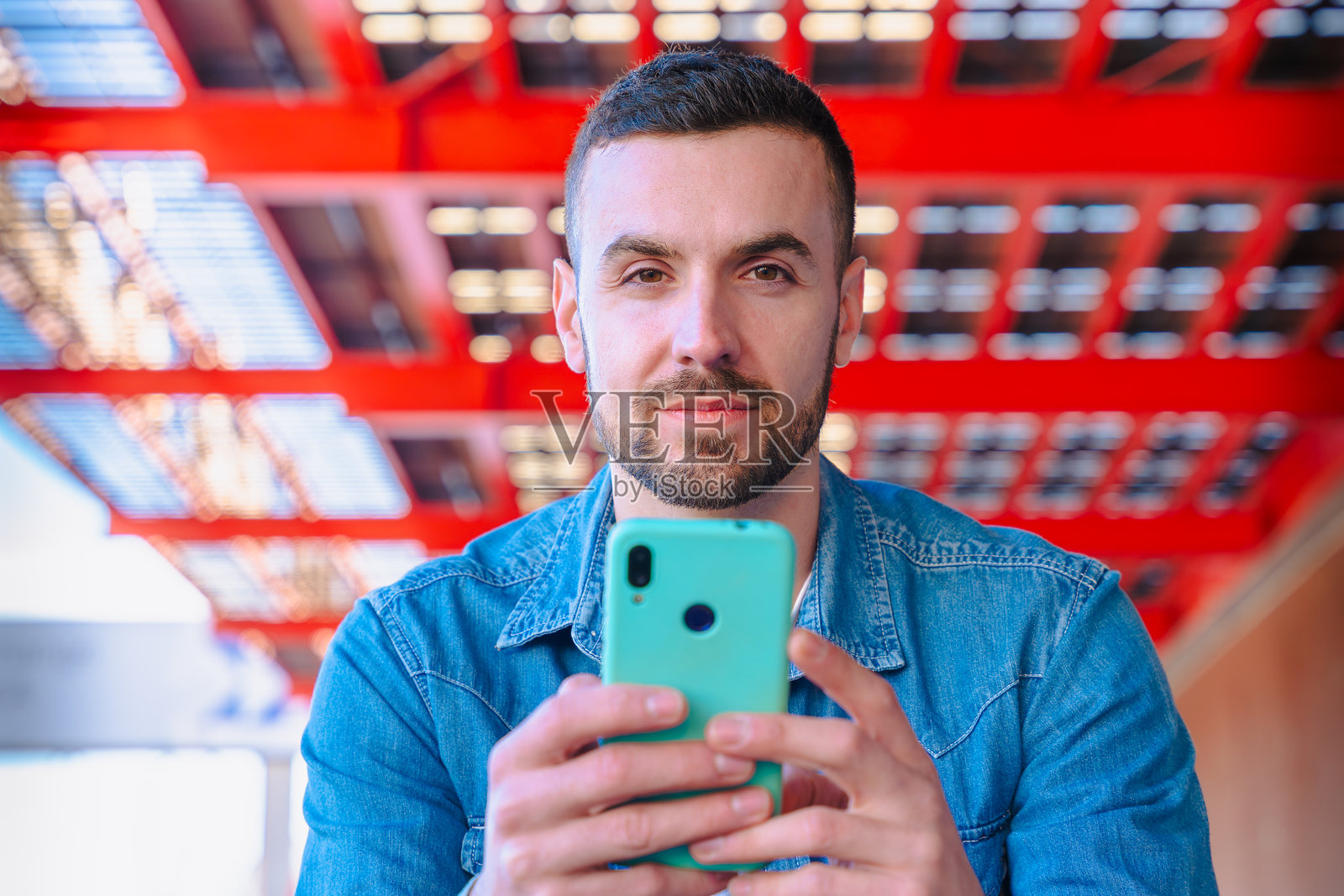 男子在红色城市背景下看着镜头，手持智能手机。照片摄影图片