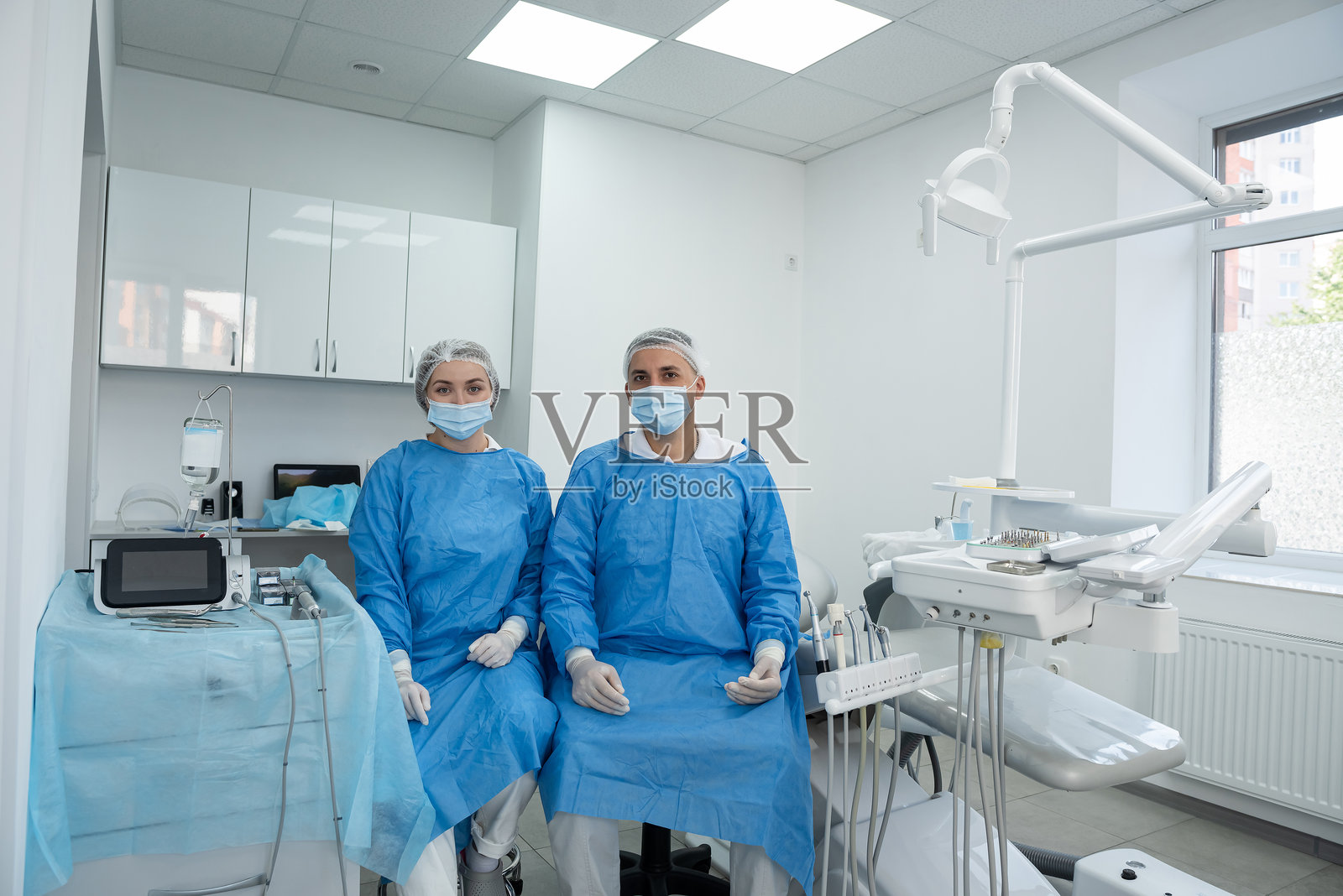 两位穿着蓝色外科大衣、戴着口罩和手套的牙医在诊所的牙科手术室内合影。团队合作照片摄影图片