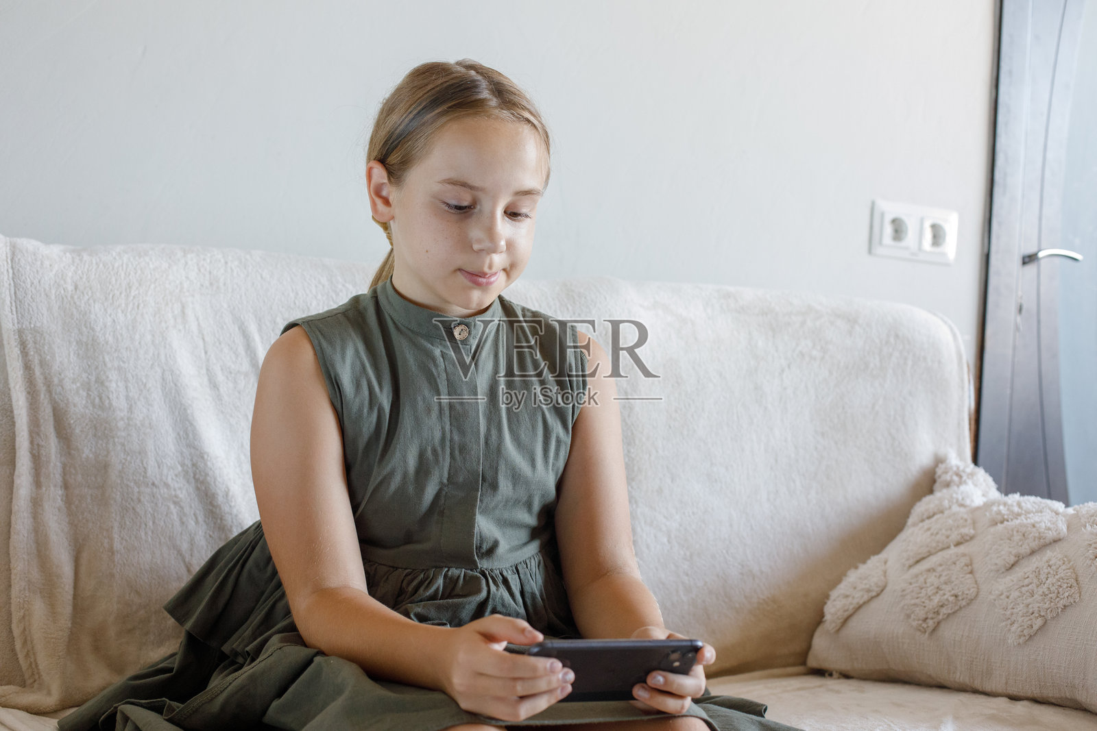 年轻女孩坐在舒适的家中沙发上使用手机阅读的肖像。儿童与智能手机。科技、沟通和休闲活动的概念。照片摄影图片