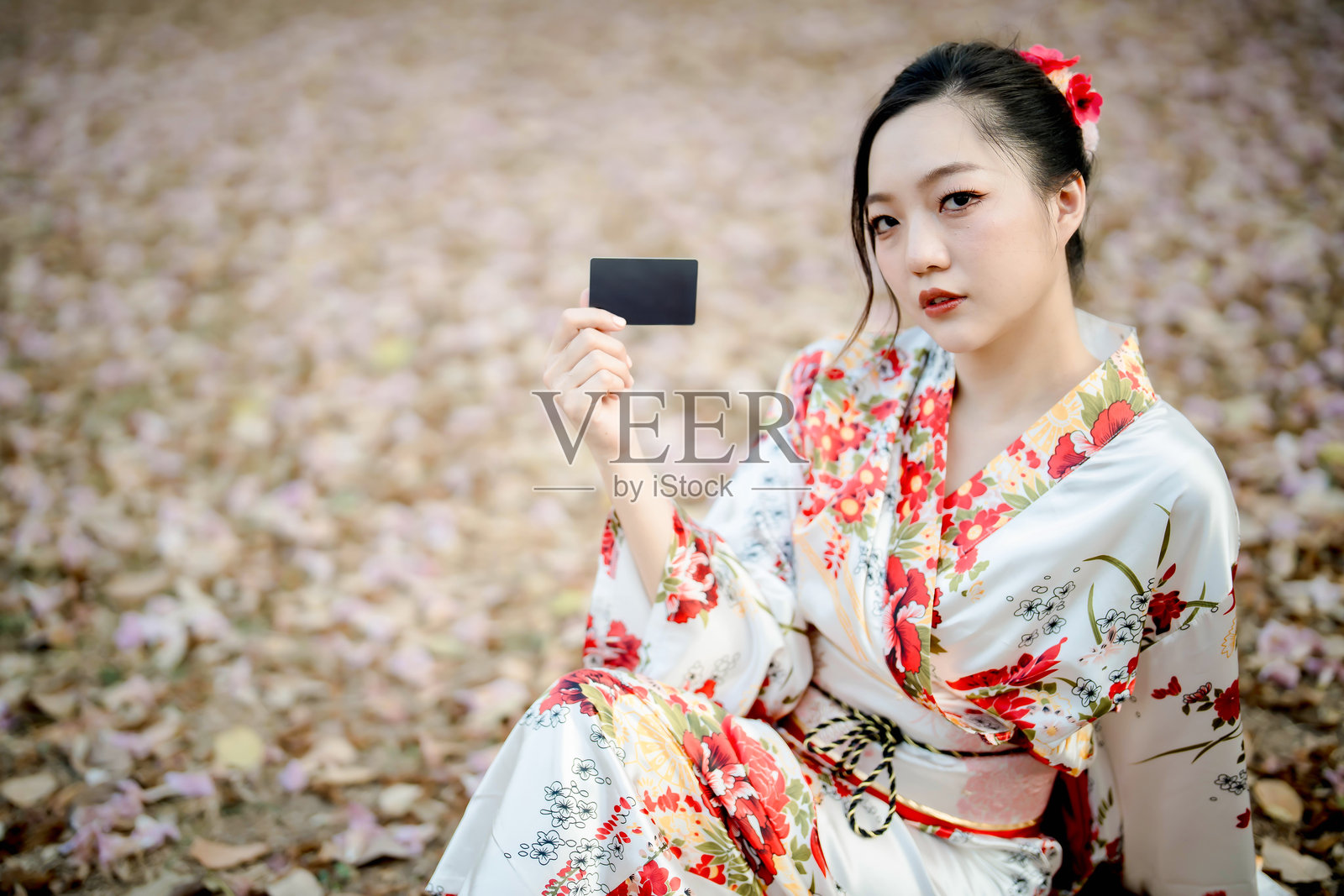 穿着传统花卉和服的美丽亚洲女性坐在秋叶地面上，手持黑色卡片，展现日本风格时尚概念照片摄影图片