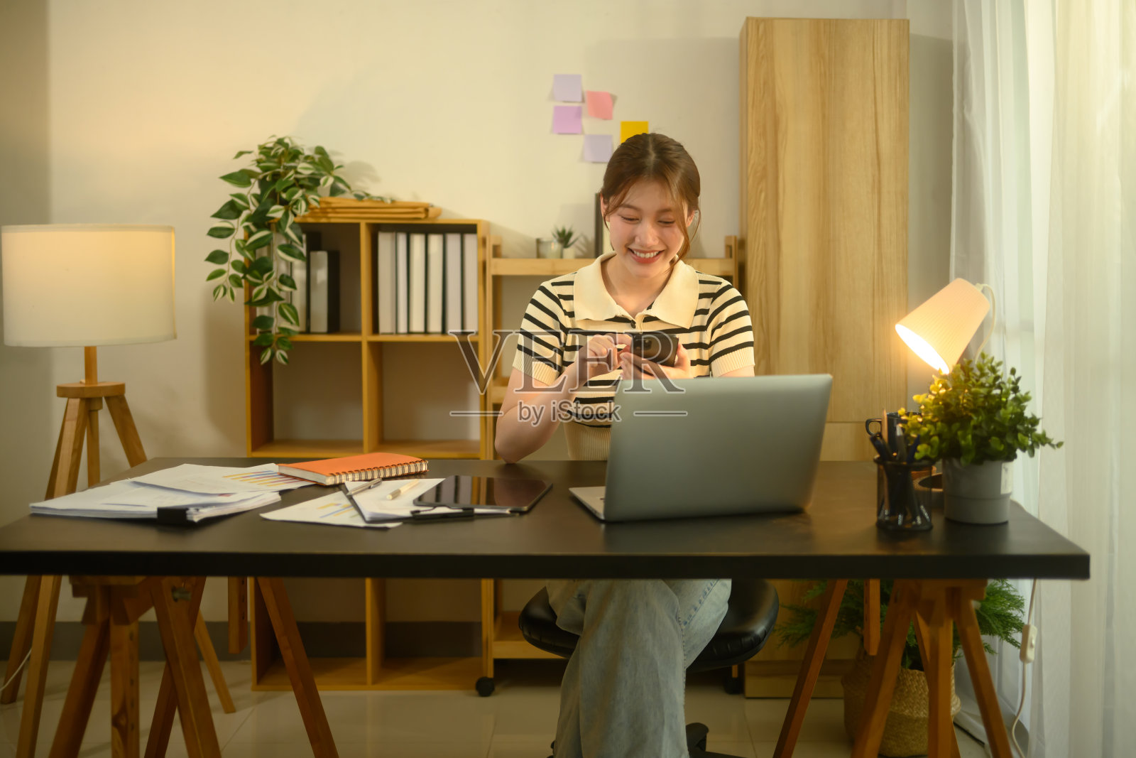 微笑的年轻女性在家办公桌上使用智能手机，同时使用笔记本电脑。照片摄影图片