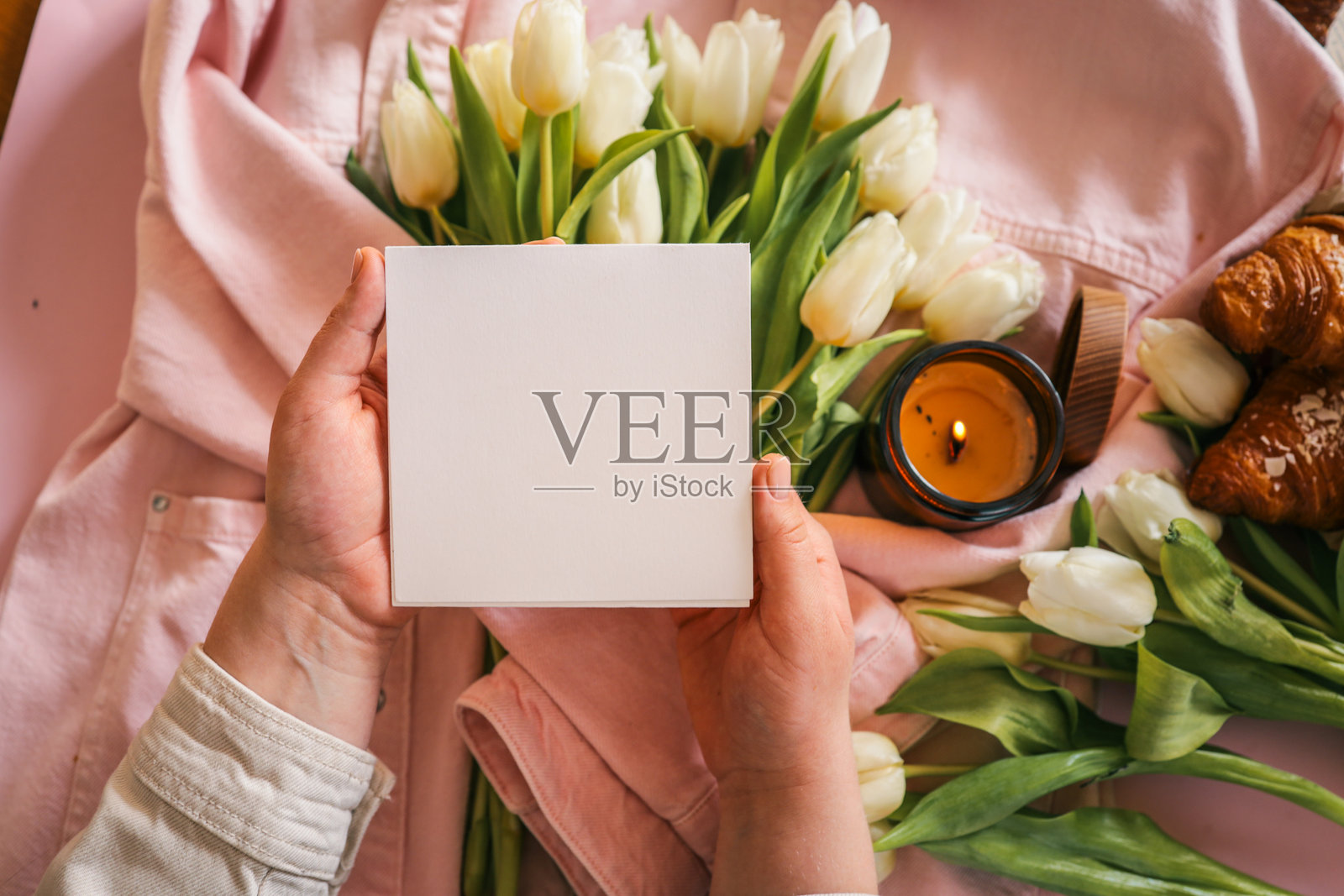 手握空白白卡片，背景是温馨的场景，白色郁金香、燃烧的蜡烛、粉色外套和可颂面包，柔和的粉色背景。非常适合用于样机或贺卡设计。照片摄影图片