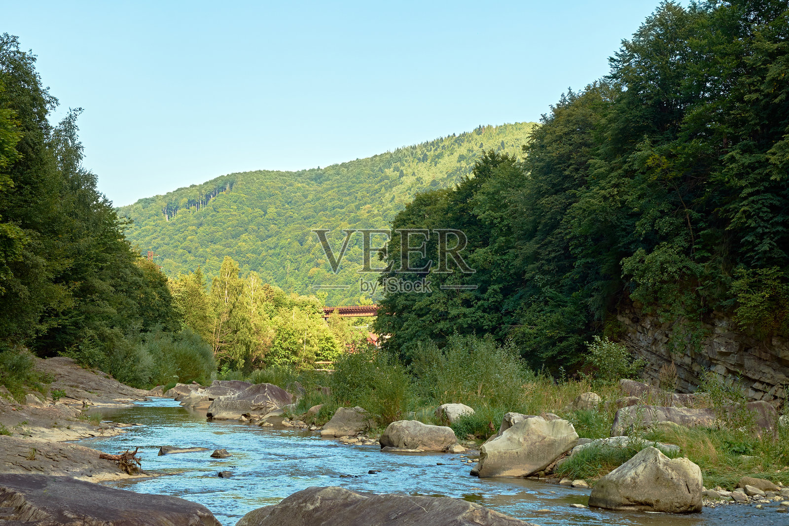 阳光明媚的夏日，风景如画的山间河流，岩石岸边绿树成荫，还有一座横跨瀑布的桥。照片摄影图片