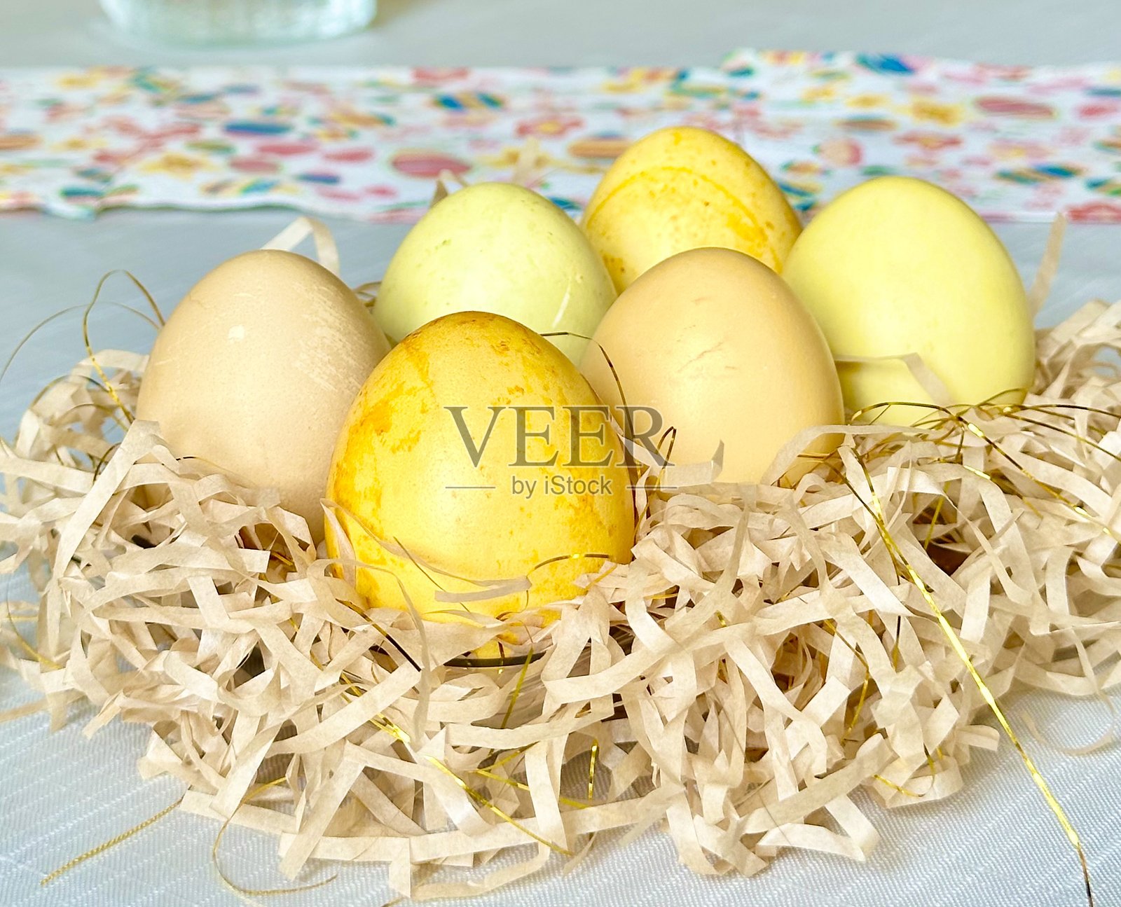 用姜黄自然染色的黄色和米色复活节彩蛋，放在带有金色丝线的 shredded paper 上，摆放在五彩斑斓的桌布上。照片摄影图片