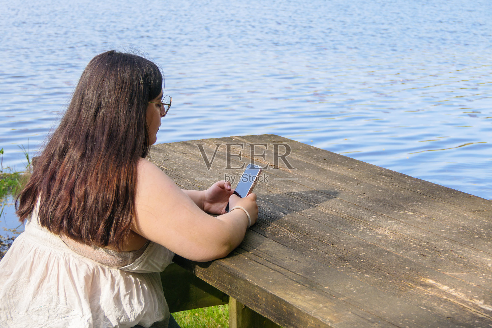 一位超重的年轻女性坐在湖边，背对着镜头使用手机。照片摄影图片