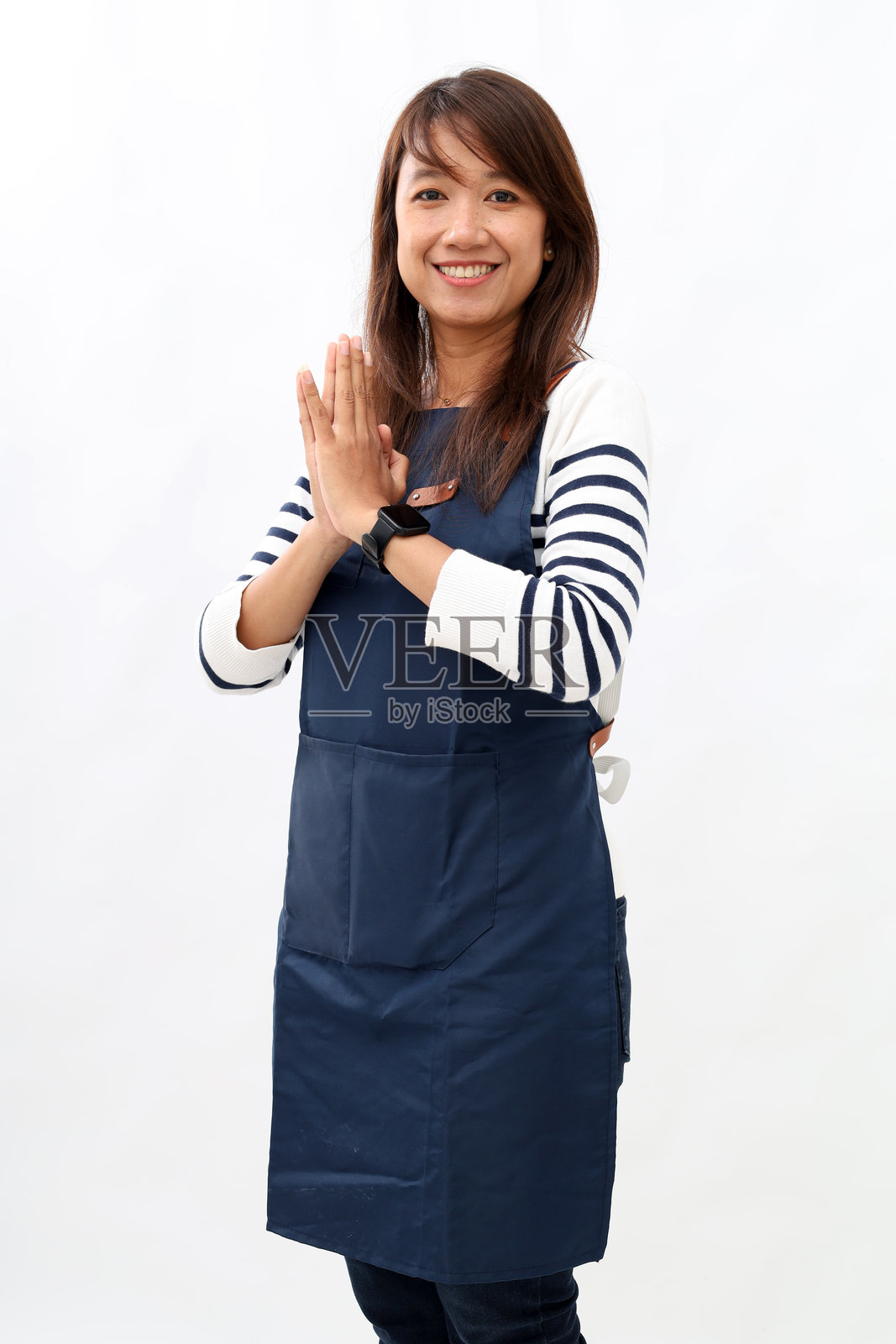 年轻的亚洲女性咖啡师穿着蓝色围裙，在咖啡店工作，双手合十，面带微笑友好地迎接顾客，背景为白色。小型企业创业概念。照片摄影图片
