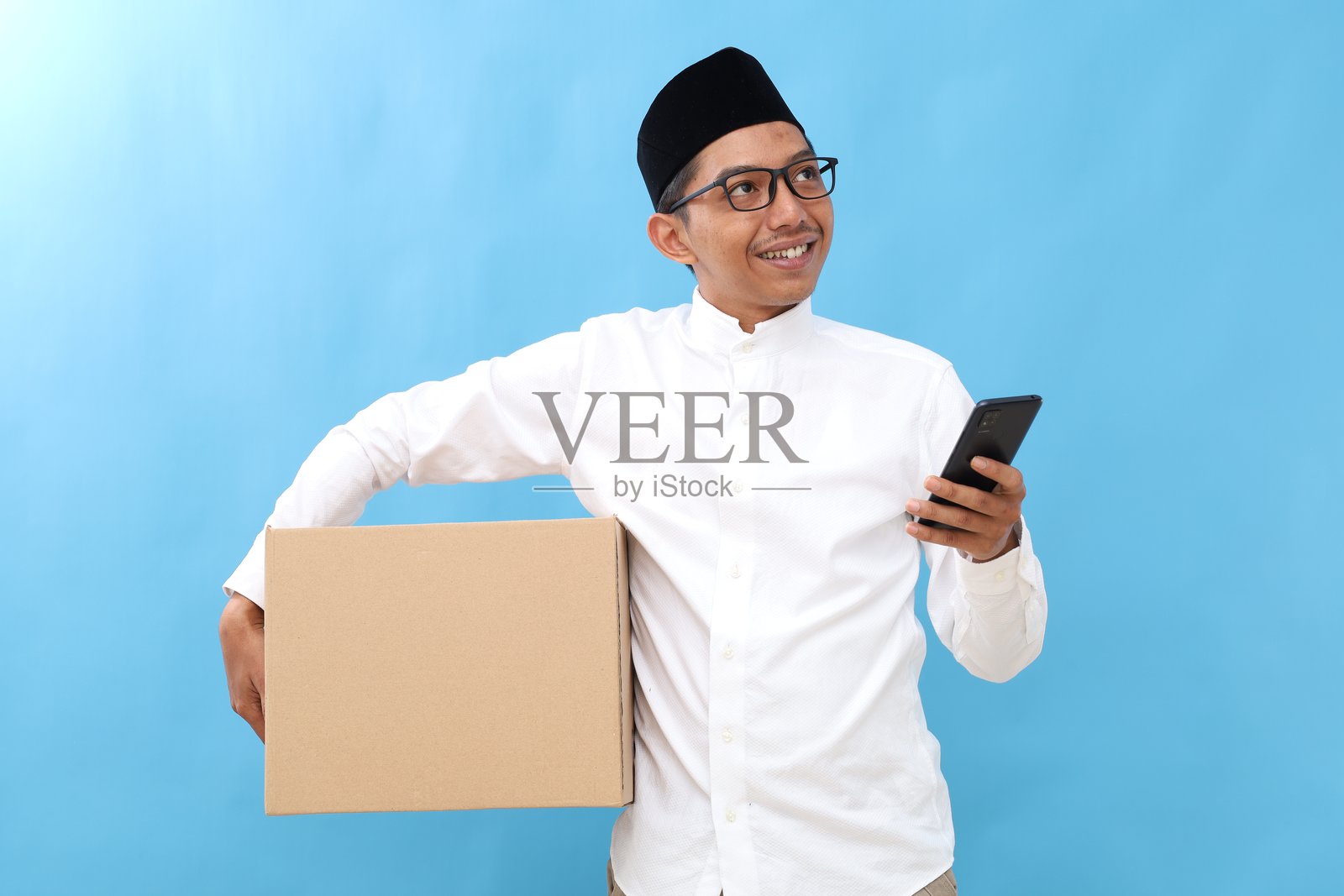 快乐的亚洲穆斯林男子一手拿着纸箱，一手拿着手机，抬头望着天空。正在回乡过拉马丹开斋节庆祝活动。照片摄影图片