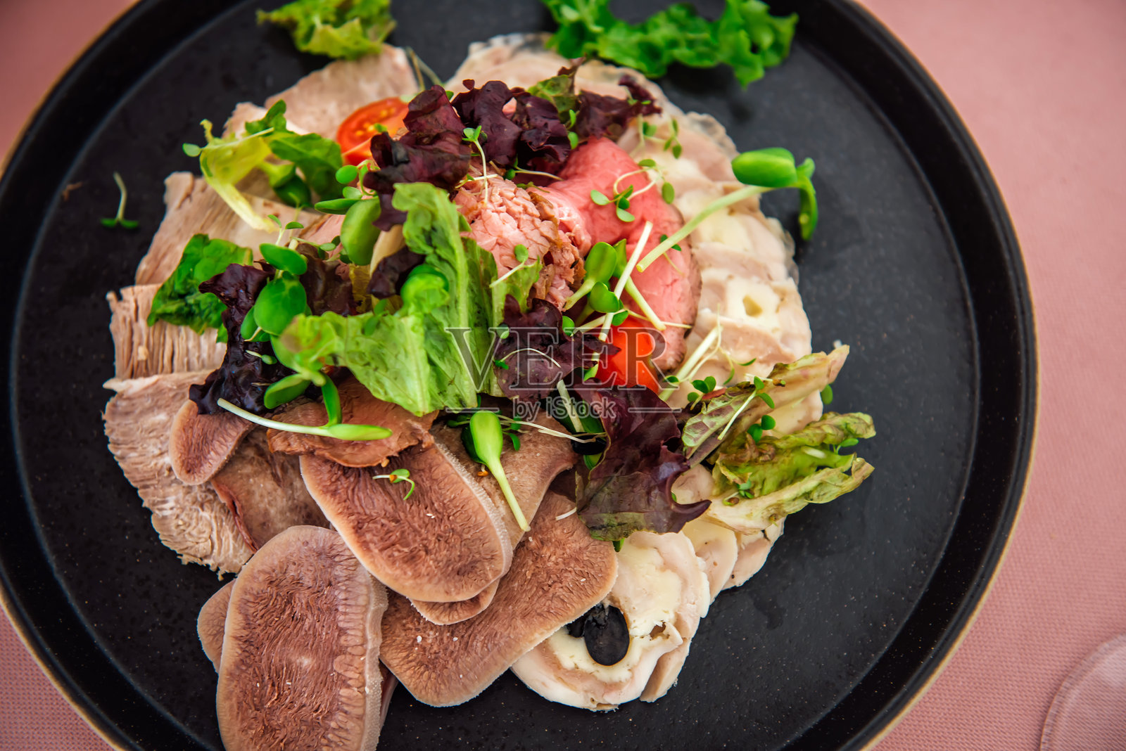 各种肉类小吃，黑盘上有煮牛舌、烤牛肉和肉卷，搭配生菜叶和微型绿植。餐厅菜肴摆放在浅粉色桌布上。照片摄影图片