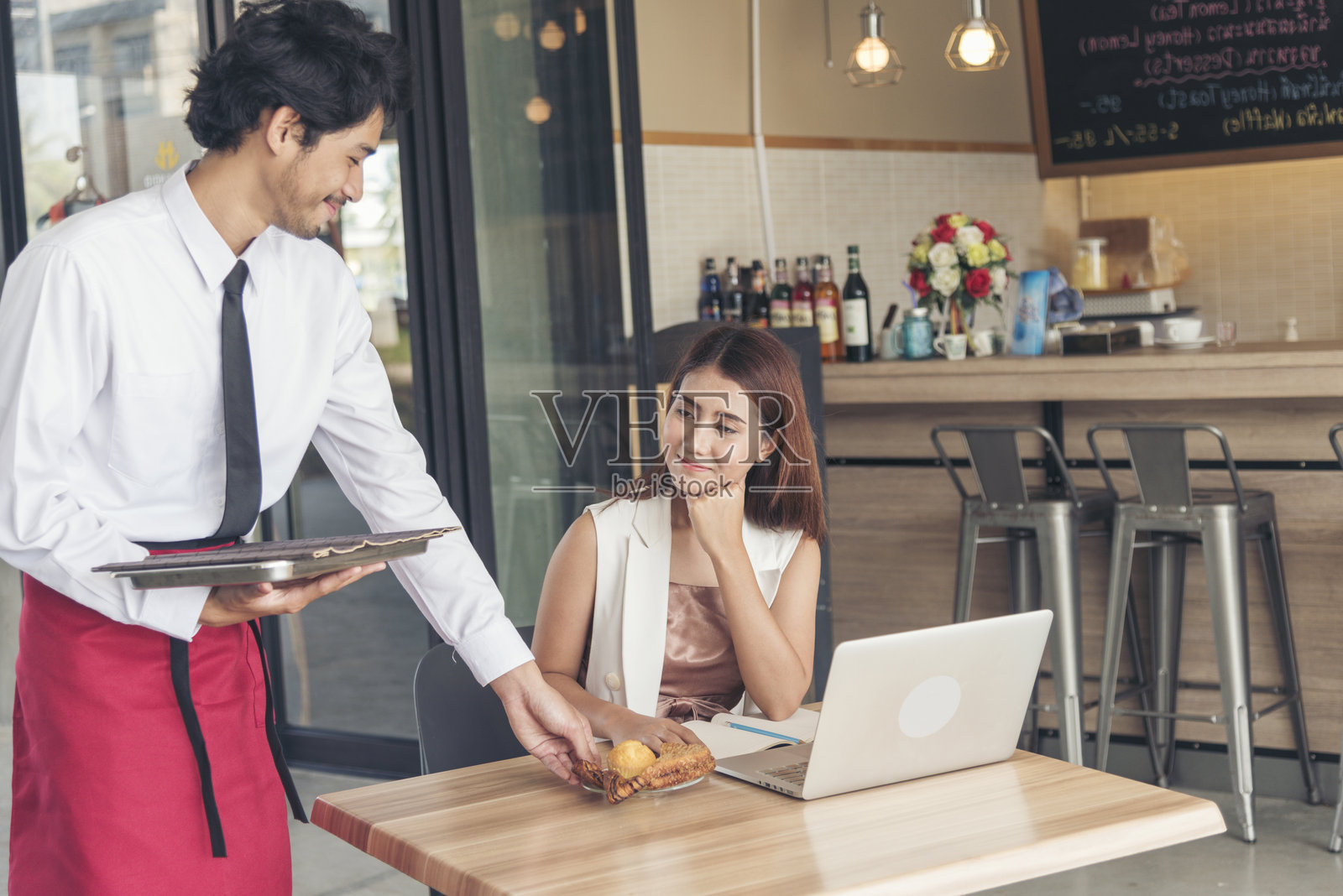 帅气的服务员微笑着为年轻美丽的女性上菜。女商人在午餐时间使用笔记本电脑工作。服务员在咖啡馆为顾客端上咖啡杯。照片摄影图片