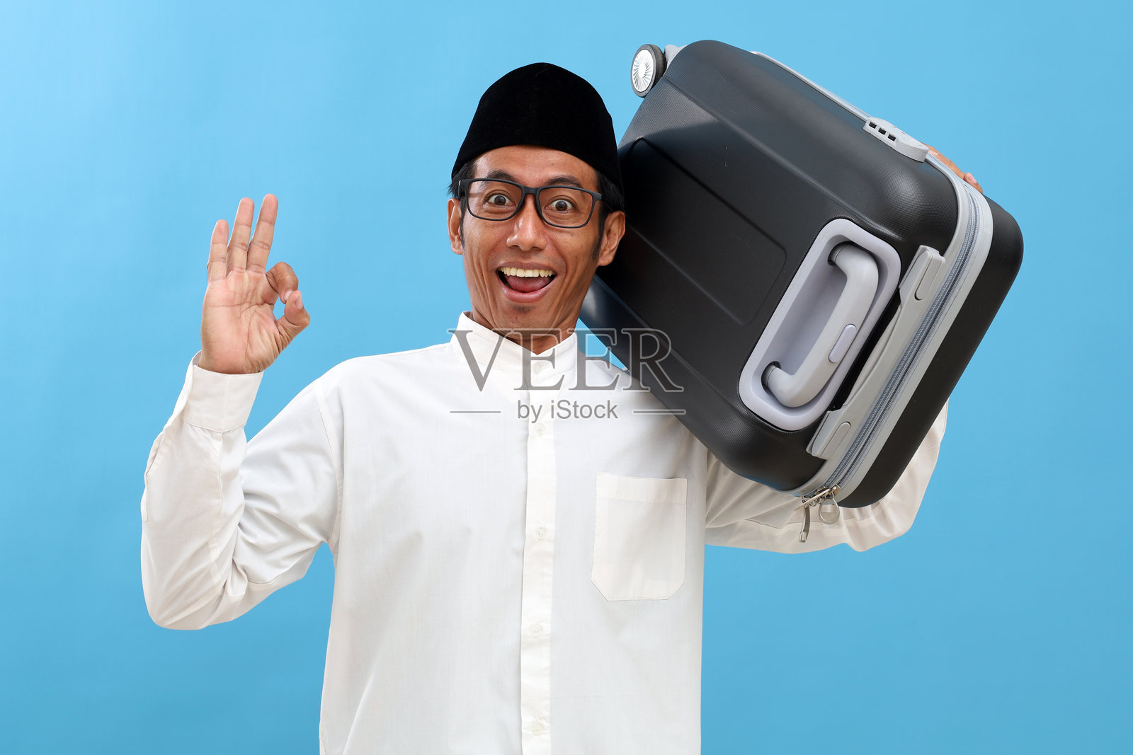 快乐的亚洲穆斯林男子提着旅行箱，做出“好的”手势。准备回乡过斋月开斋节庆祝活动。照片摄影图片