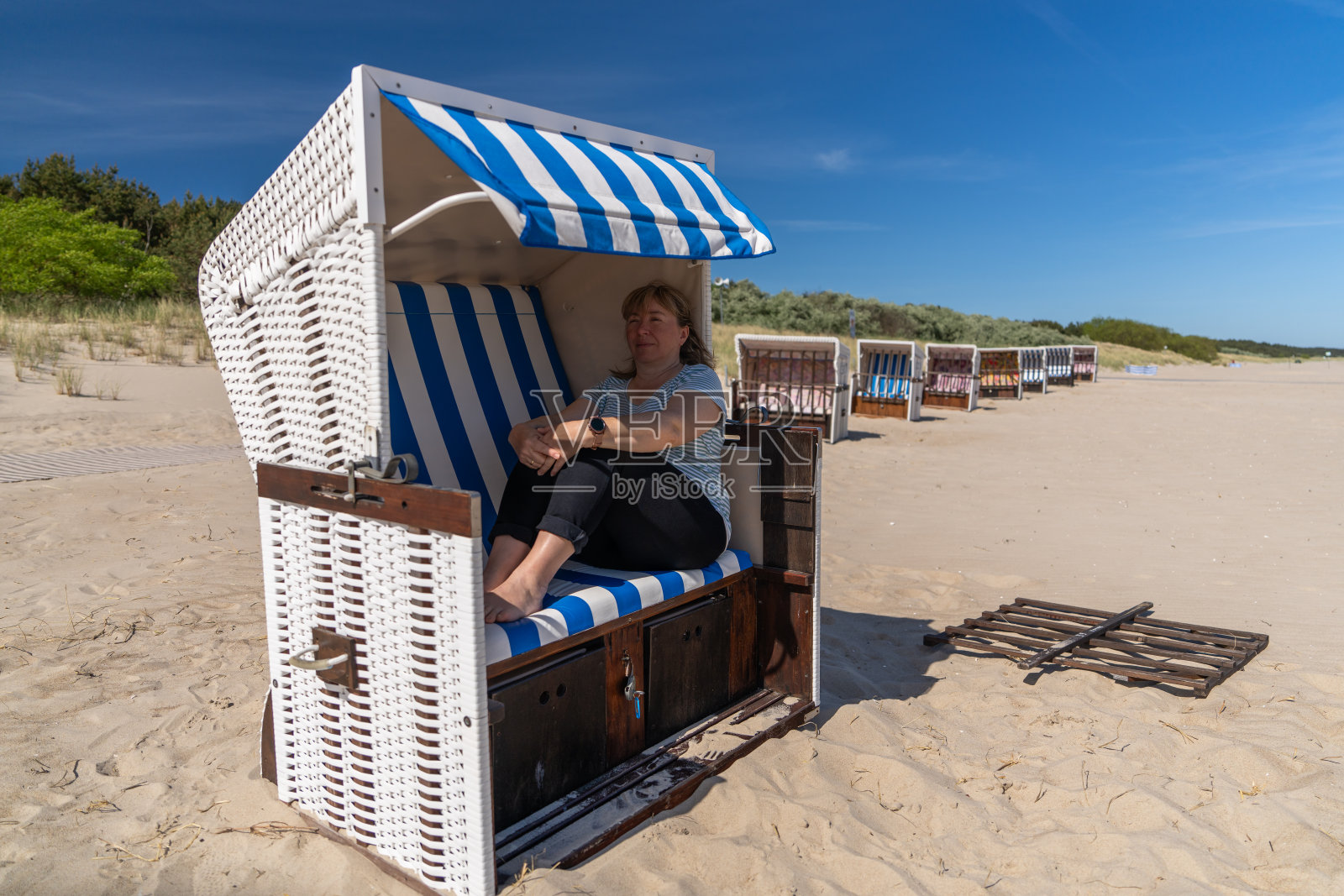 波罗的海的夏季假期。身穿白色长袍和米色帽子的欧洲美女坐在沙滩上的躺椅上，阳光明媚的日子里阅读书籍，白色沙滩上放松。侧面视图。照片摄影图片