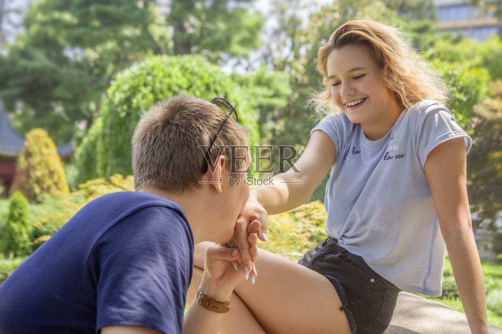 男孩亲吻一位微笑快乐女孩的手。她的手指上戴着订婚戒指。阳光明媚的夏日，公园散步。照片摄影图片