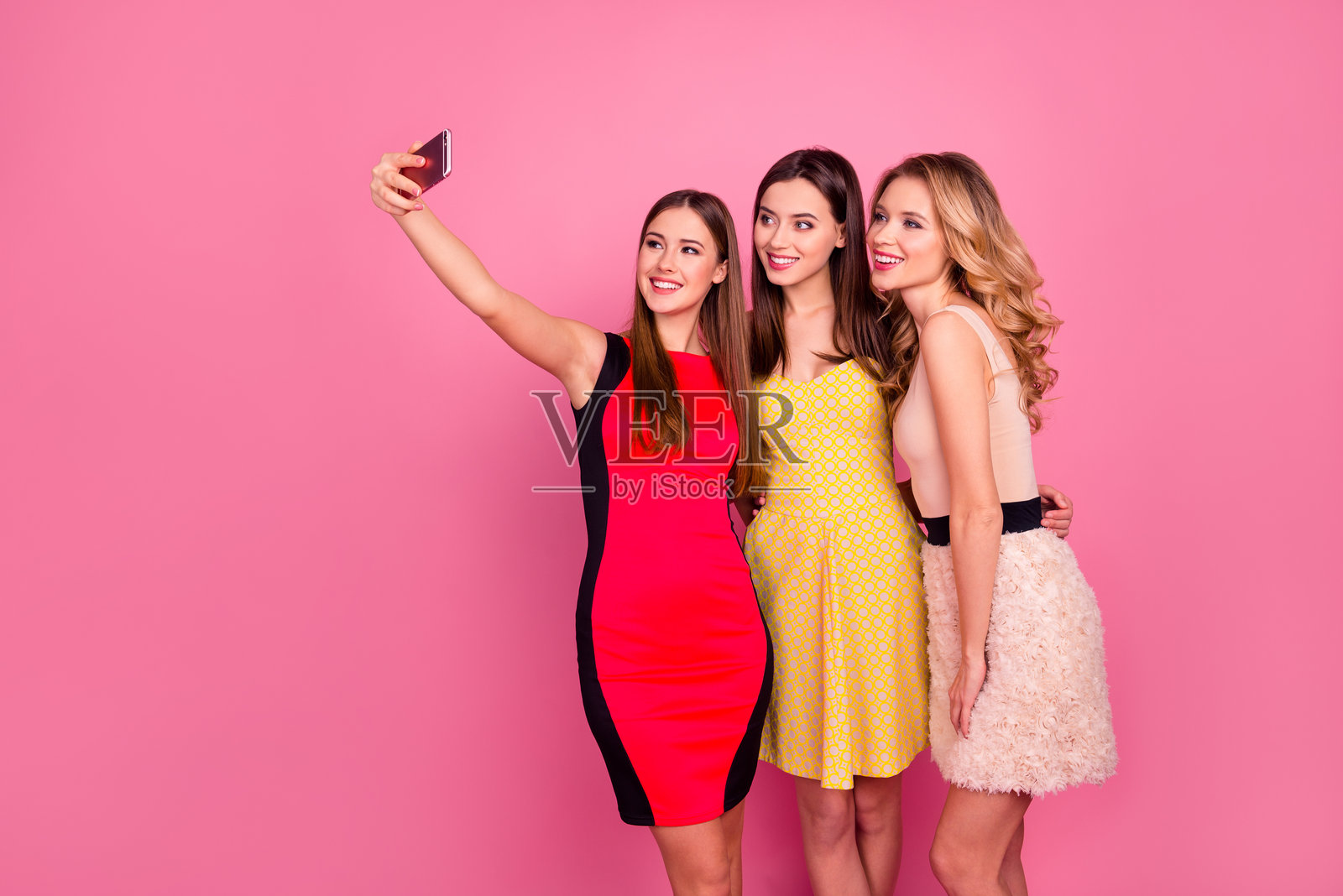 三位漂亮女孩穿着鸡尾酒裙在粉色背景下拥抱自拍照片摄影图片