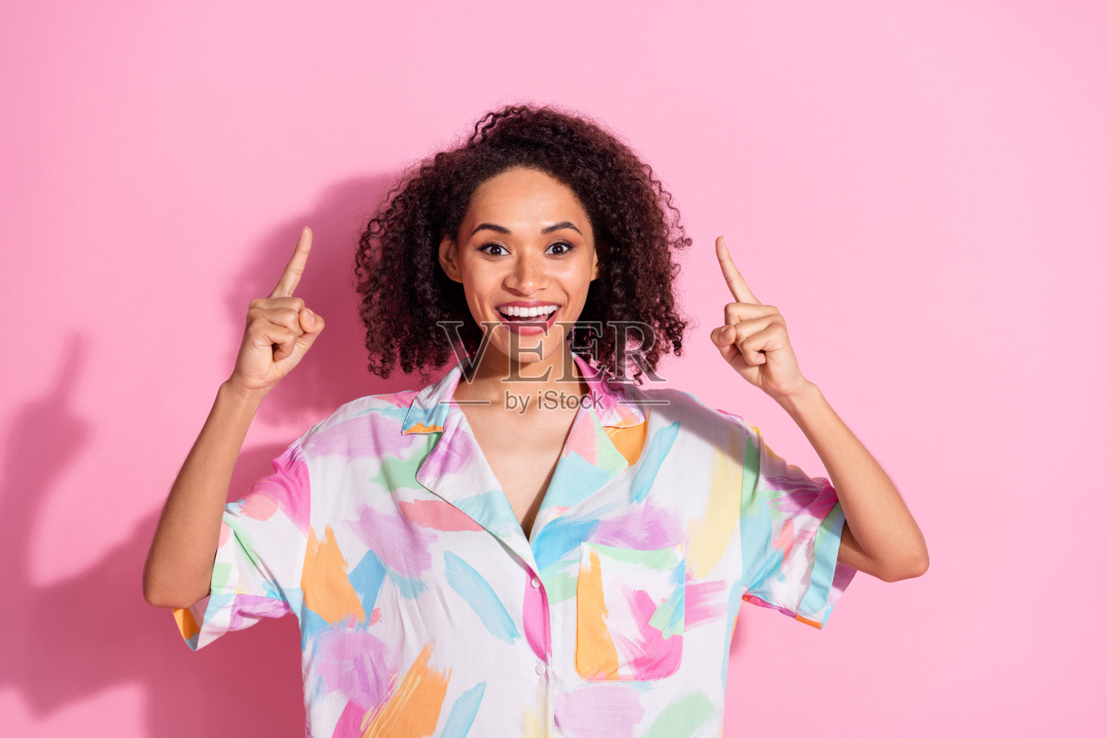 穿着鲜艳印花衬衫的快乐年轻女性在粉色背景前做出俏皮的手势，展现出自信和积极的态度。照片摄影图片
