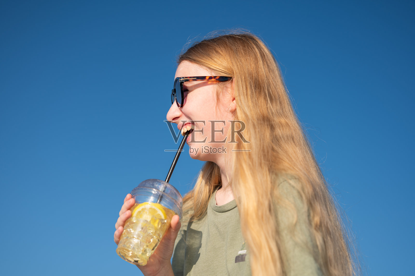 年轻女性戴着太阳镜，享受着清爽的饮料，背景是蓝天。照片摄影图片