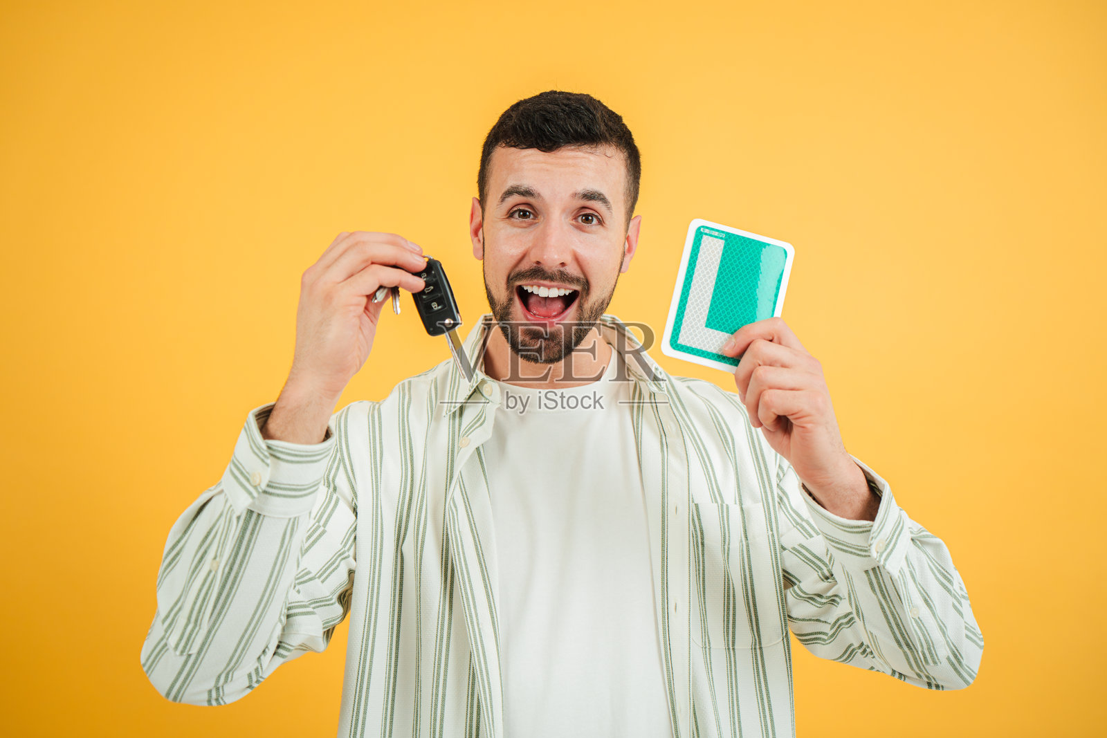 年轻的白人举着新的驾驶执照L牌和车钥匙，面带灿烂的笑容，因通过驾驶学校考试而感到开心和自豪，面向镜头，背景为黄色。照片摄影图片