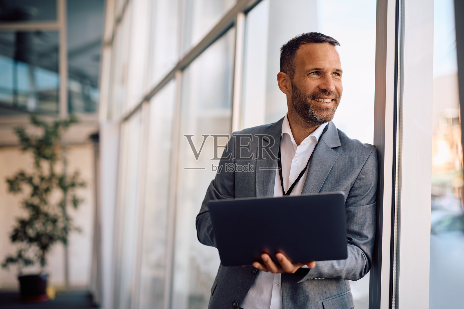 快乐的商人在办公室使用笔记本电脑，同时透过窗户看外面。照片摄影图片