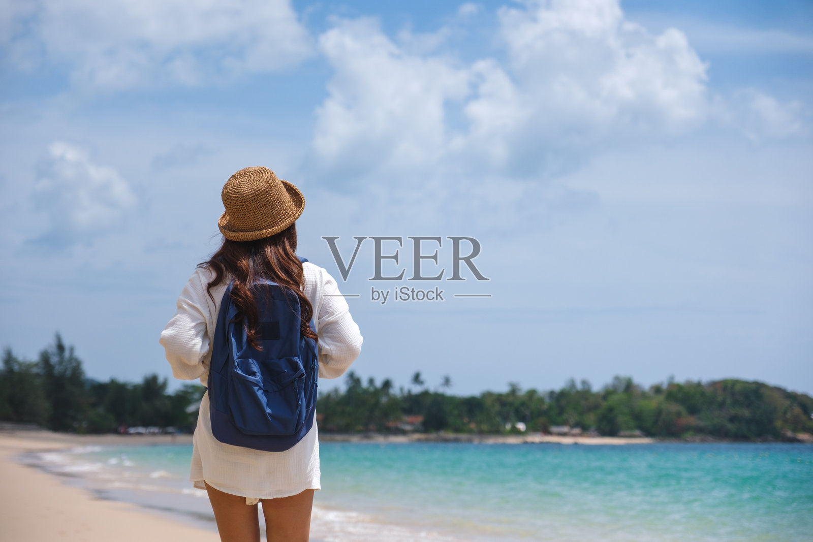 背影的女性旅行者，戴着帽子和背包，站在海边的沙滩上照片摄影图片