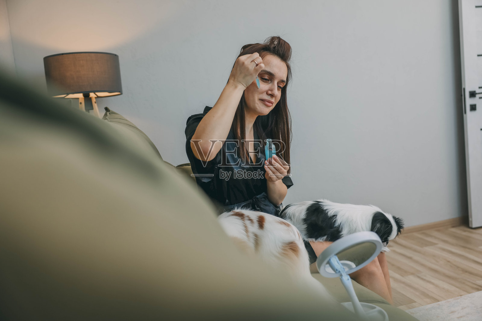 女性在宁静的家居环境中进行美容护肤，坐在绿色沙发上涂抹面部精华，身边有两只小狗陪伴。照片摄影图片