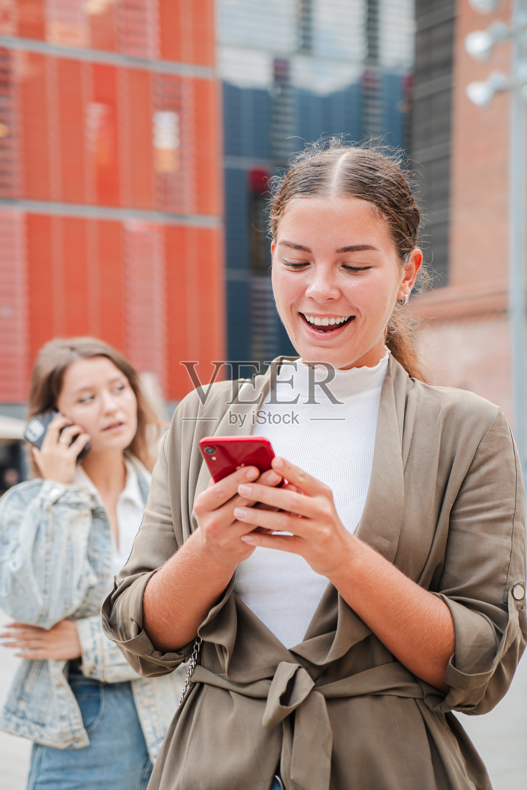 垂直构图。快乐的年轻女性正在用智能手机发短信，旁边另一位女性正在打手机。这一迷人的瞬间突显了朋友之间的智能手机沟通。照片摄影图片