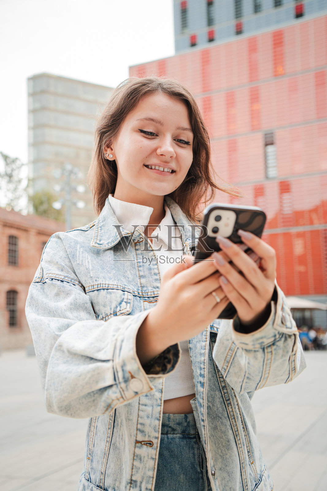 垂直构图。一个微笑的少女在户外使用手机，穿着牛仔夹克，站在红色建筑前，享受与智能手机互动的轻松时刻。照片摄影图片