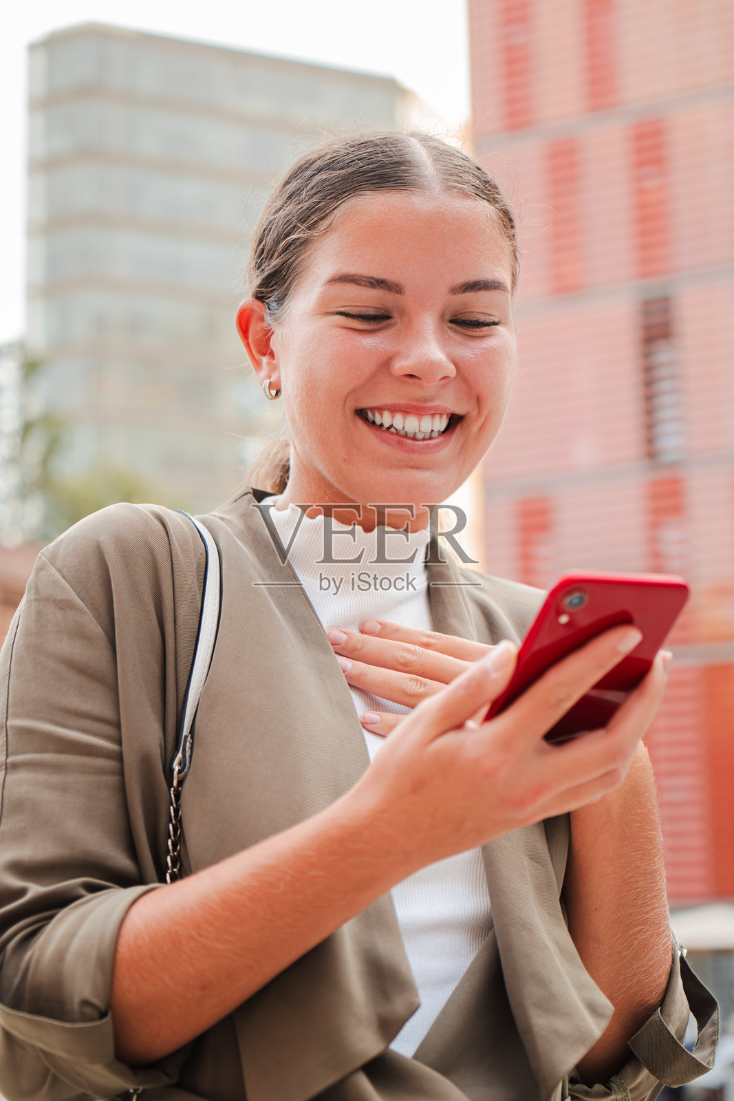 年轻女性在户外微笑着使用手机，查看信息或社交媒体，背景为城市环境。她穿着休闲商务服装，手持手机，背后是现代建筑。照片摄影图片