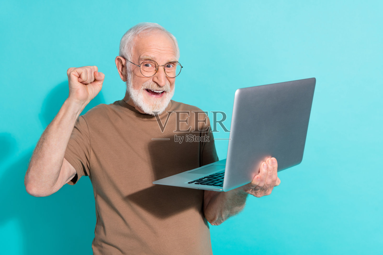 吸引人的快乐灰发男士在明亮的蓝色背景下使用笔记本电脑庆祝加密货币。照片摄影图片
