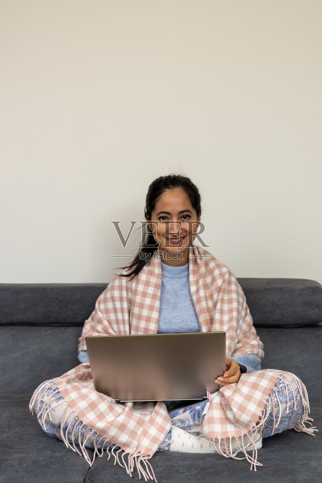 女性自由职业者在家远程工作，盘腿坐在沙发上，裹着柔软的毛毯，笔记本电脑打开，微笑自信地看着镜头，明亮的客厅内饰。照片摄影图片