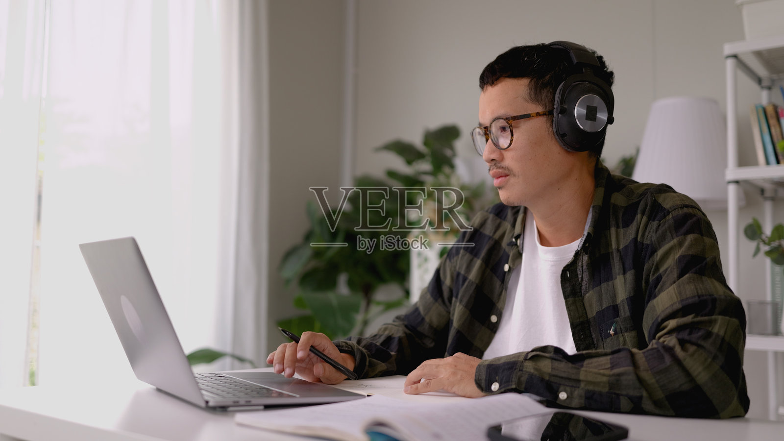 亚洲男子在家办公室里戴着耳机听音乐并在笔记本电脑上工作。照片摄影图片