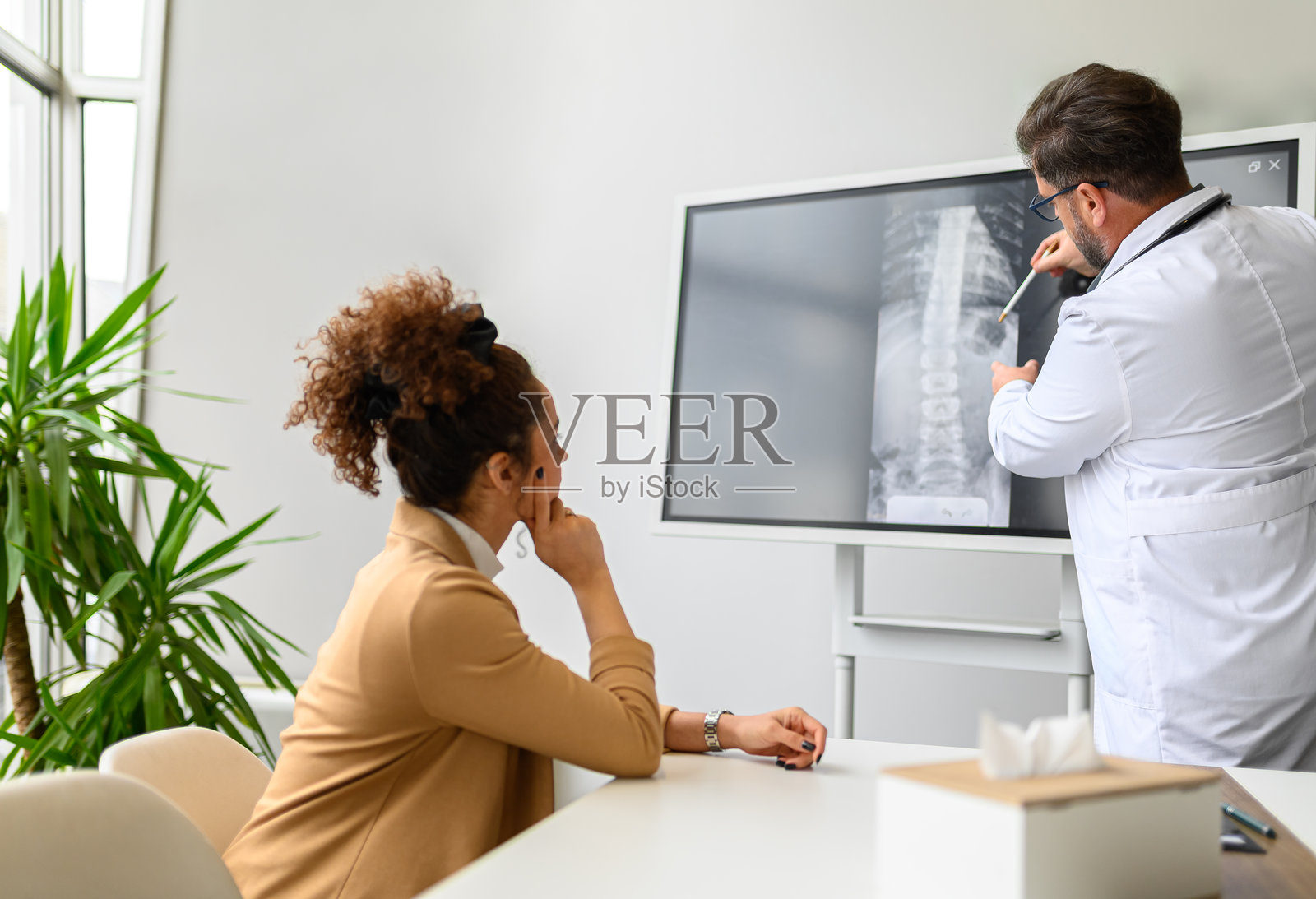 男医生在医院向女性患者展示并解释脊柱X光片，电脑屏幕上显示相关信息。照片摄影图片