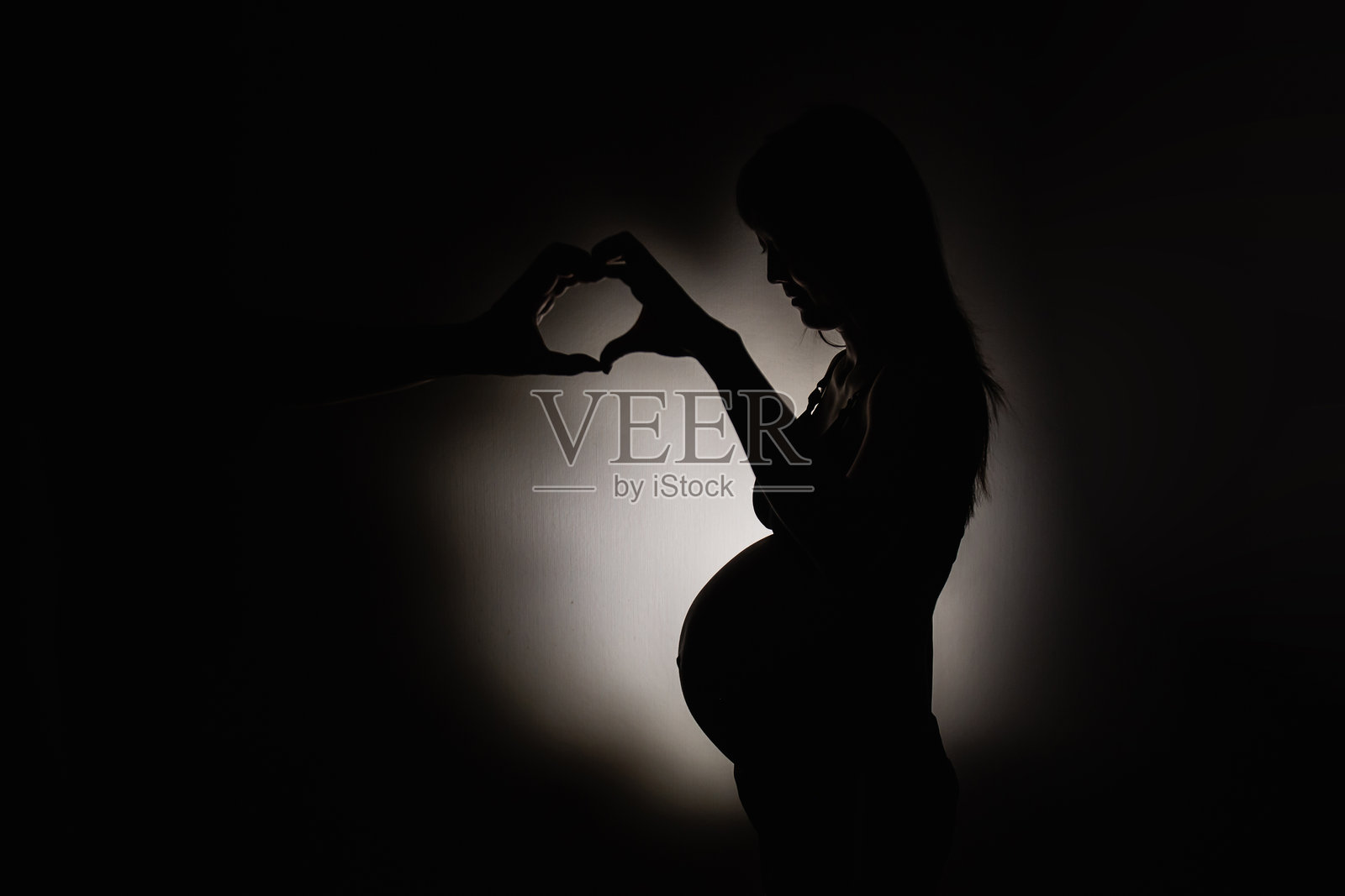 怀孕女性的剪影，双手在中心形成心形，背景中有她的父亲。概念摄影。照片摄影图片