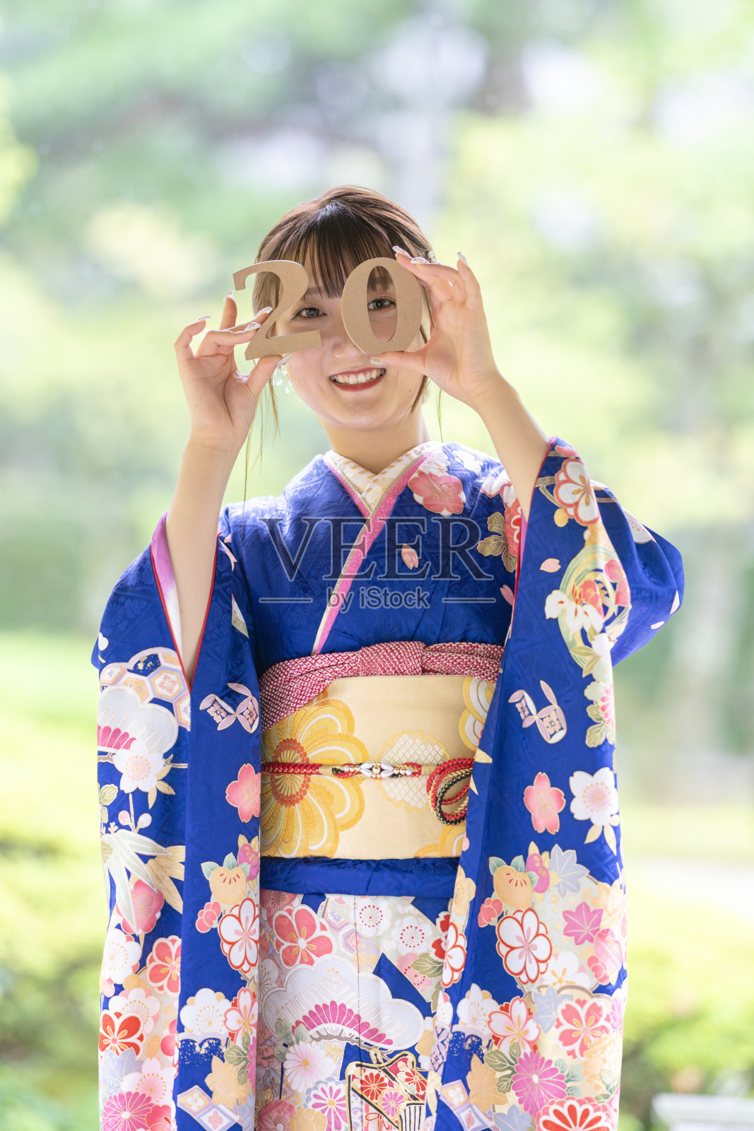 日本某城市的雨天夏日。一位穿着蓝色青少年和服的日本女性在传统花园中。她手中拿着数字2和0，笑着庆祝20岁生日。照片摄影图片