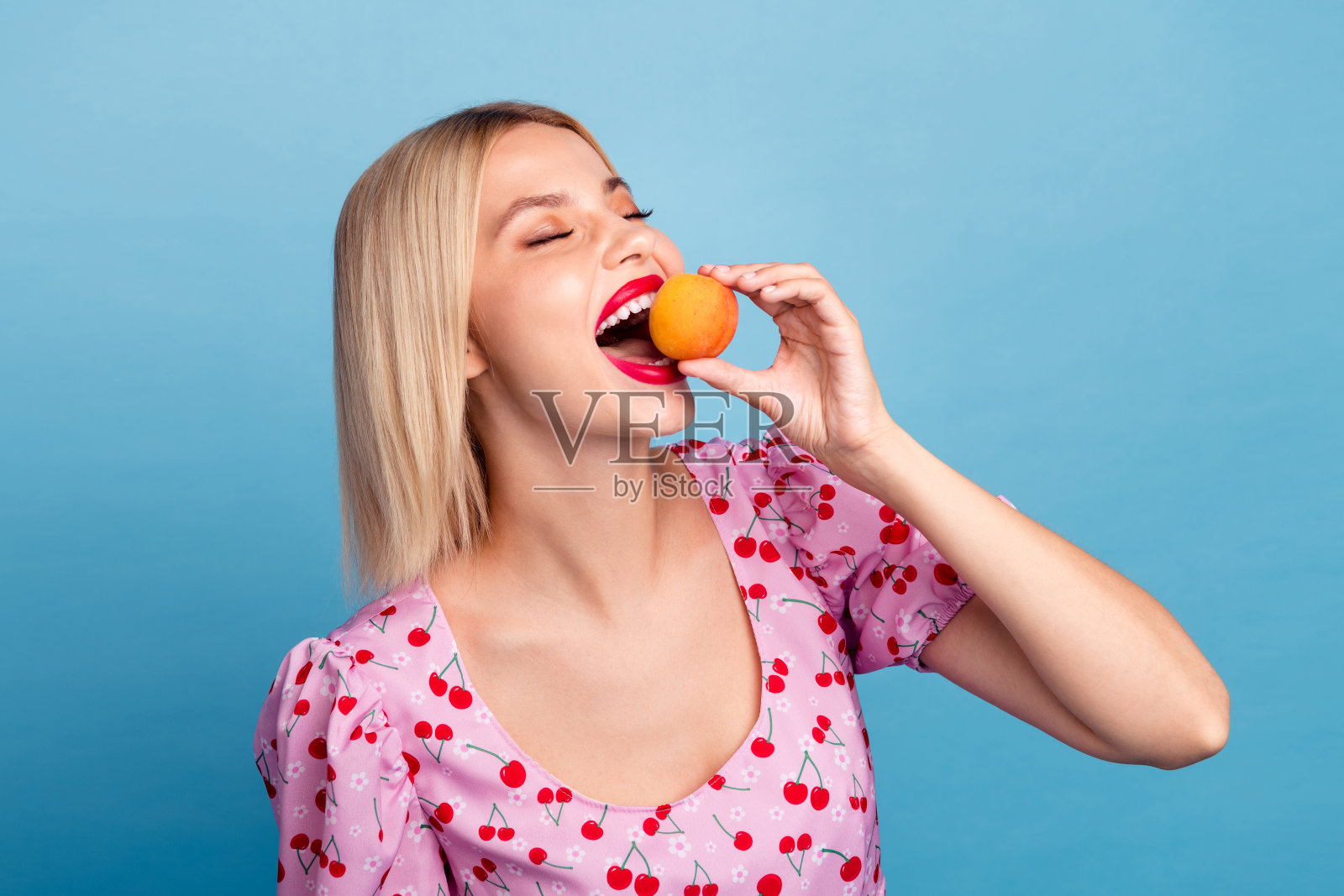 快乐的年轻女性，金色头发，享受着新鲜的杏子，背景是明亮的蓝色，穿着迷人的樱桃图案上衣。照片摄影图片