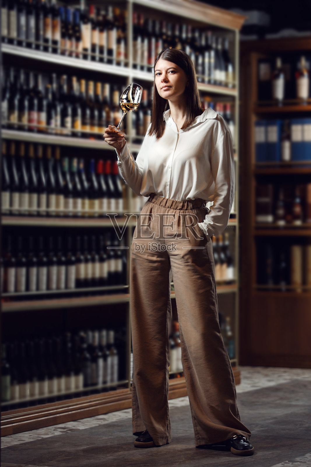 熟练的女性酒庄专家在酒窖环境中品尝酒，周围是酒瓶架。照片摄影图片