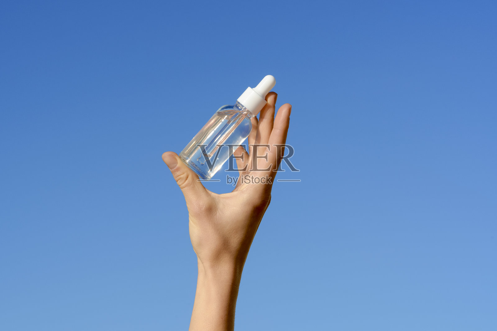 女性手握着一个透明的玻璃瓶，瓶内装有精华液，配有滴管，背景是蓝天。美丽与美学产品的概念，护肤保湿化妆品。照片摄影图片