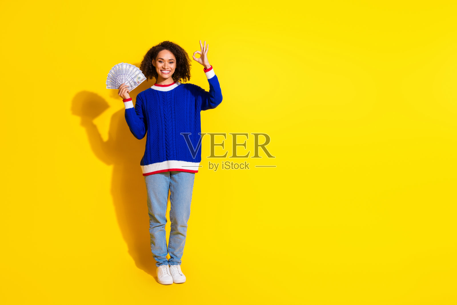 穿着休闲服的快乐女性手持钞票扇，做出顽皮的和平手势，背景为鲜艳的黄色。照片摄影图片