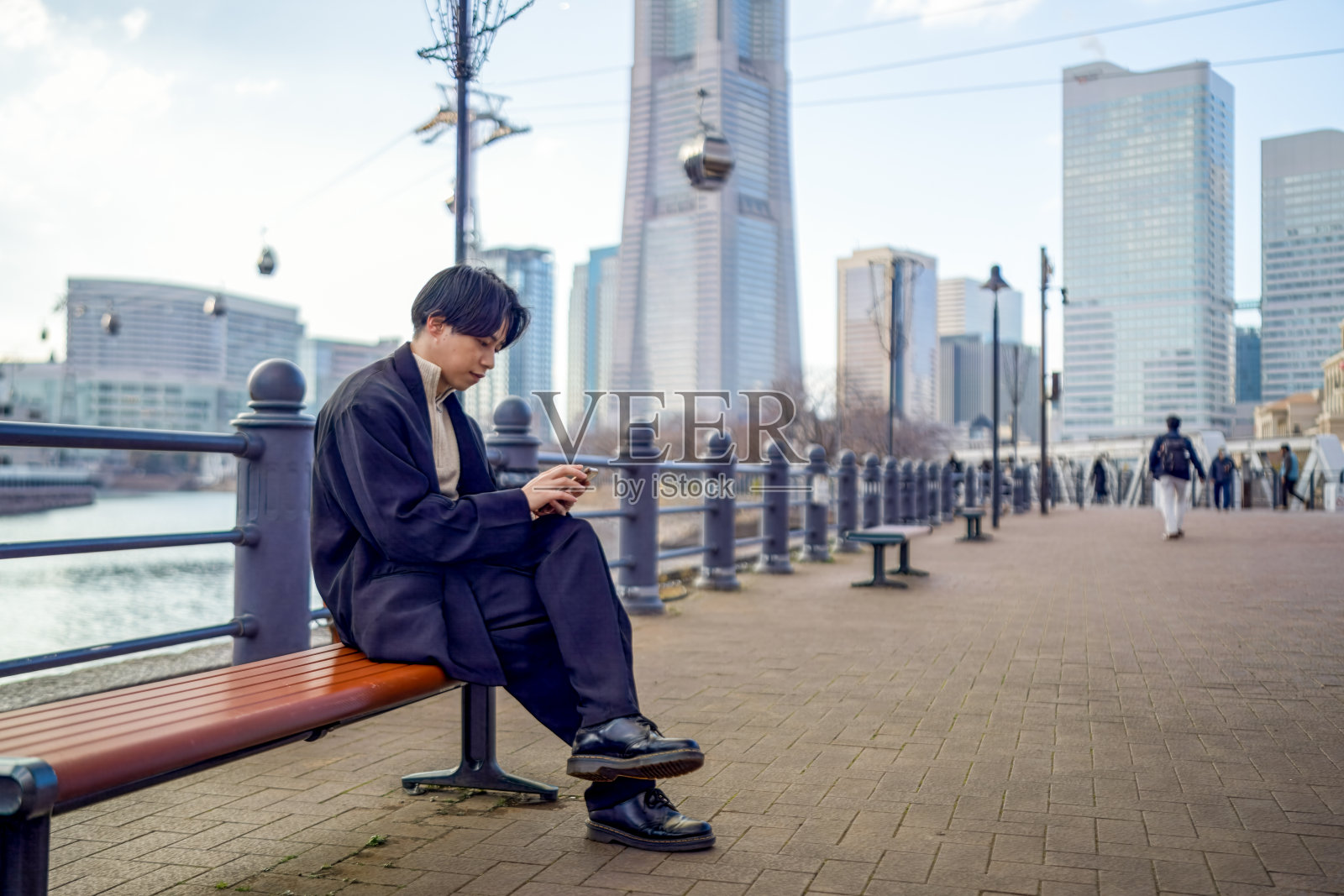 一位二十多岁的日本男子穿着黑色夹克，坐在横滨的港未来长椅上，身后是高楼大厦和现代化的天际线，他正在使用智能手机。照片摄影图片