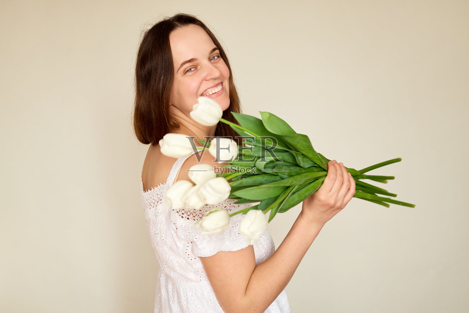 一位长着棕色长发的年轻女性愉快地微笑着，手中捧着一束白色郁金香。她站在室内，背景明亮，散发着幸福和魅力。照片摄影图片