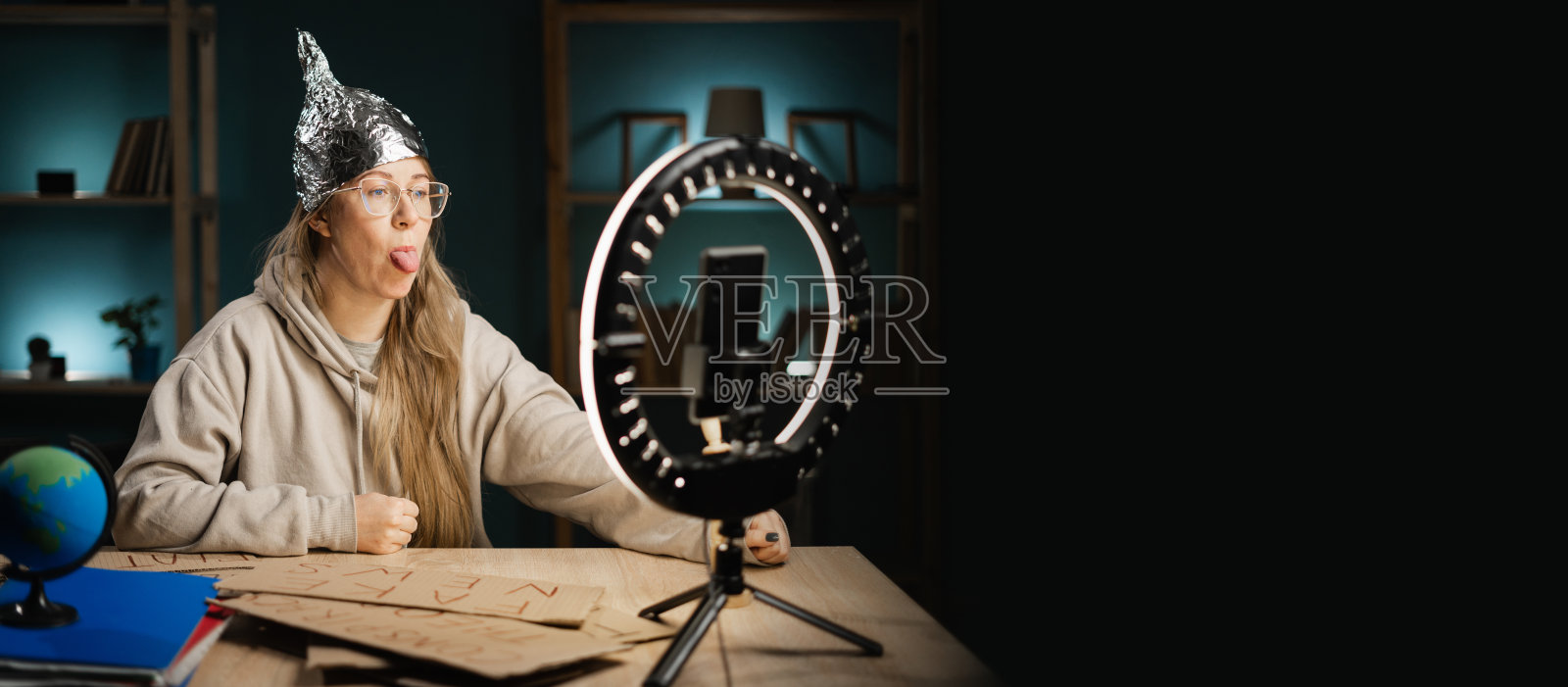 戴眼镜的疯狂阴谋论者博主在家中使用智能手机和环形灯进行直播，调皮地伸出舌头。横幅。照片摄影图片