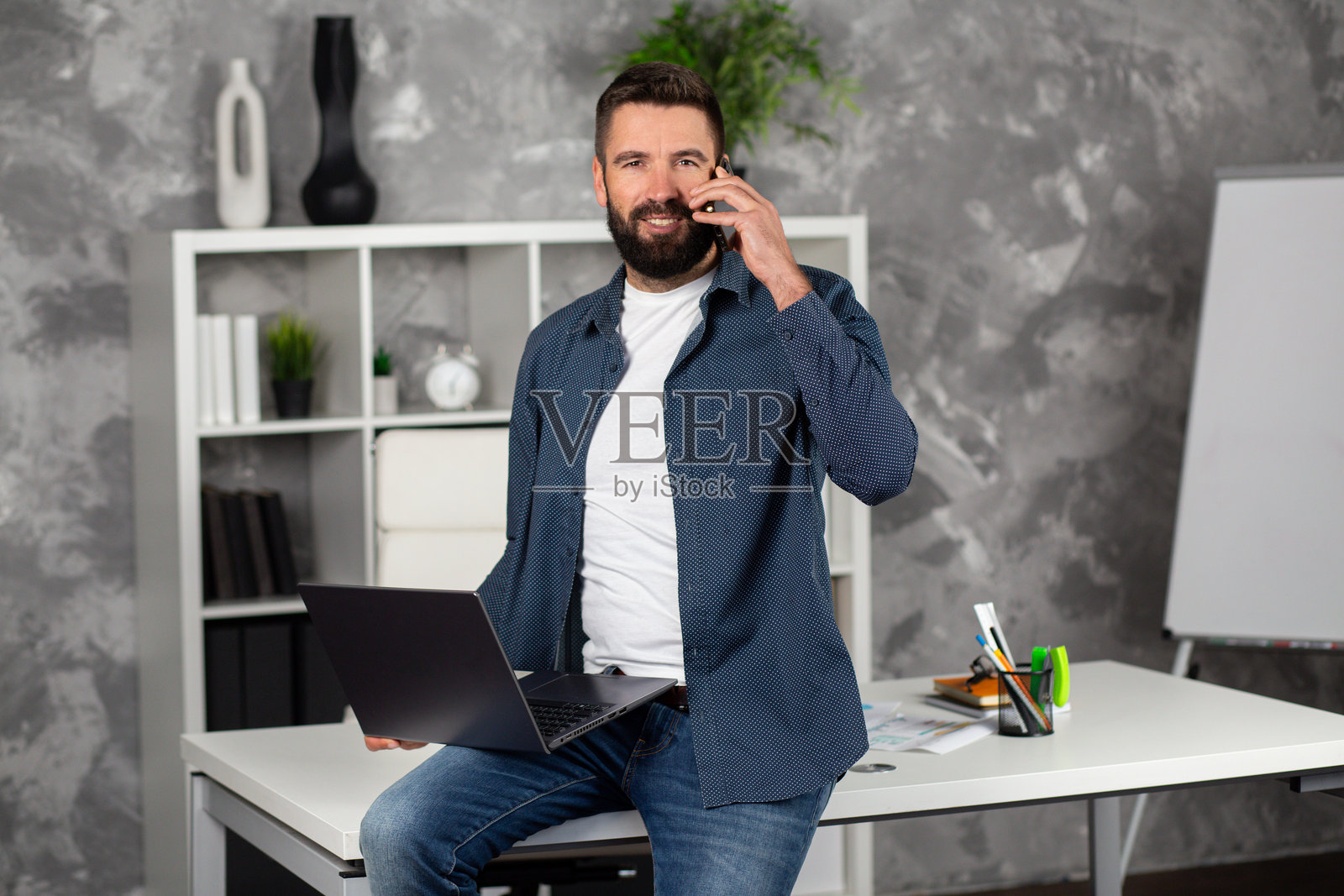 坐在办公室桌子前的帅气男子，留着胡须，手持笔记本电脑，正在打电话，面带微笑看向镜头。照片摄影图片
