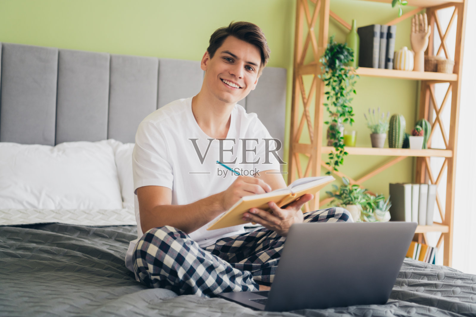 年轻男子穿着睡衣在床上写笔记，面带微笑，身处一个舒适明亮的卧室，周围环绕着植物和现代装饰。照片摄影图片