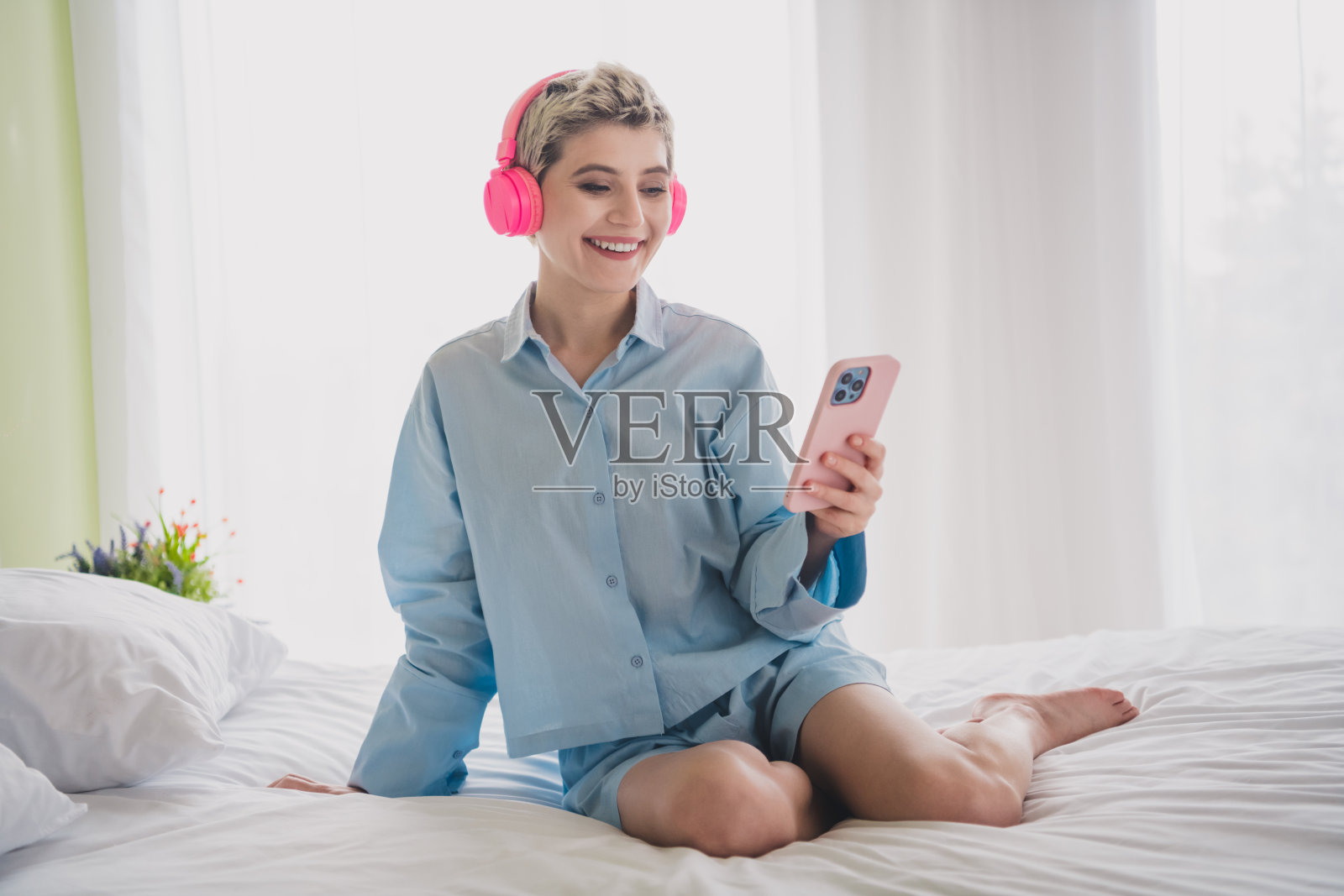 年轻女性穿着睡衣，微笑着戴着耳机听音乐，同时手里拿着智能手机。照片摄影图片