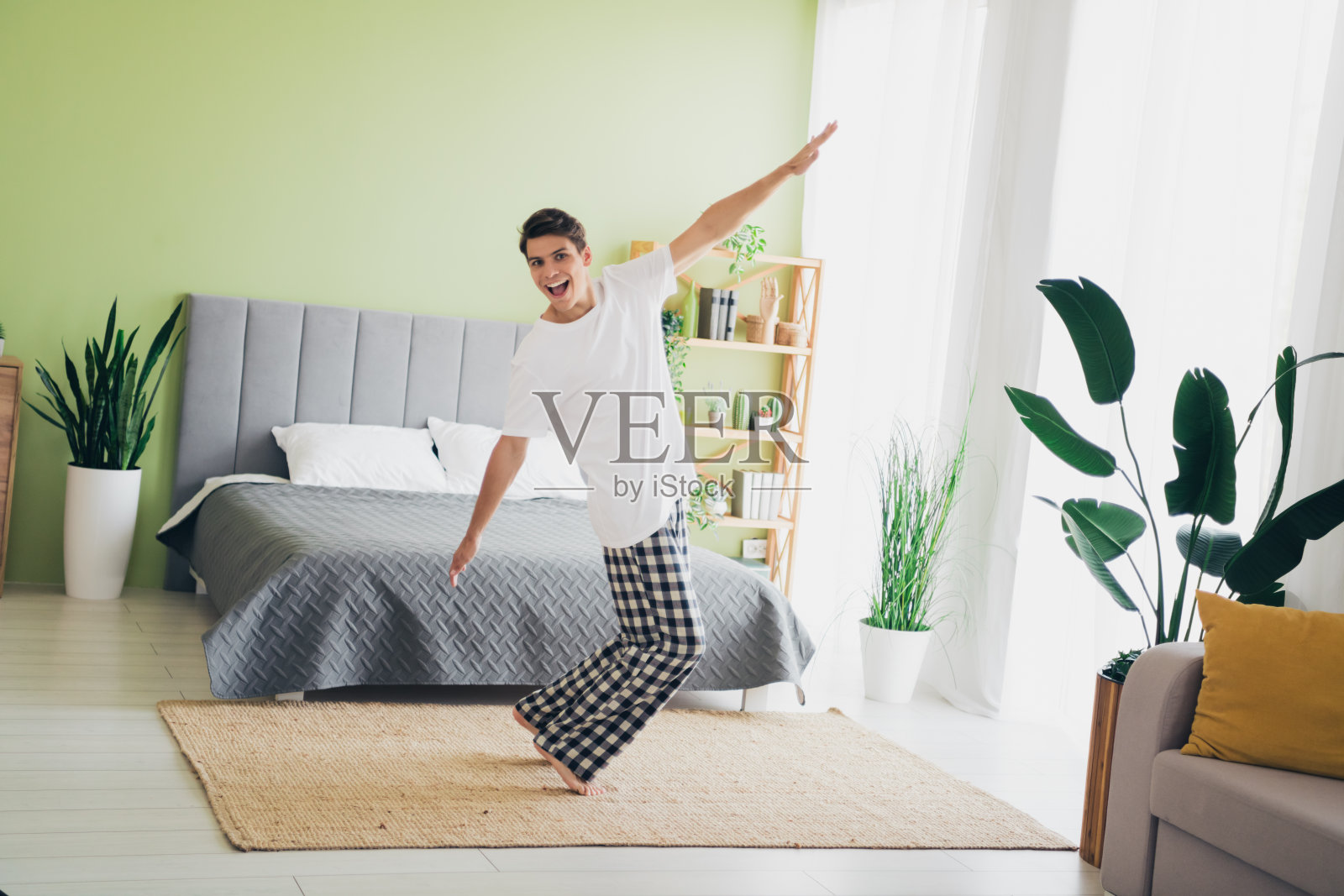 充满活力的年轻男子穿着睡衣，在明亮的现代卧室里跳舞，房间装饰着绿色，家具舒适，晨光洒入。照片摄影图片