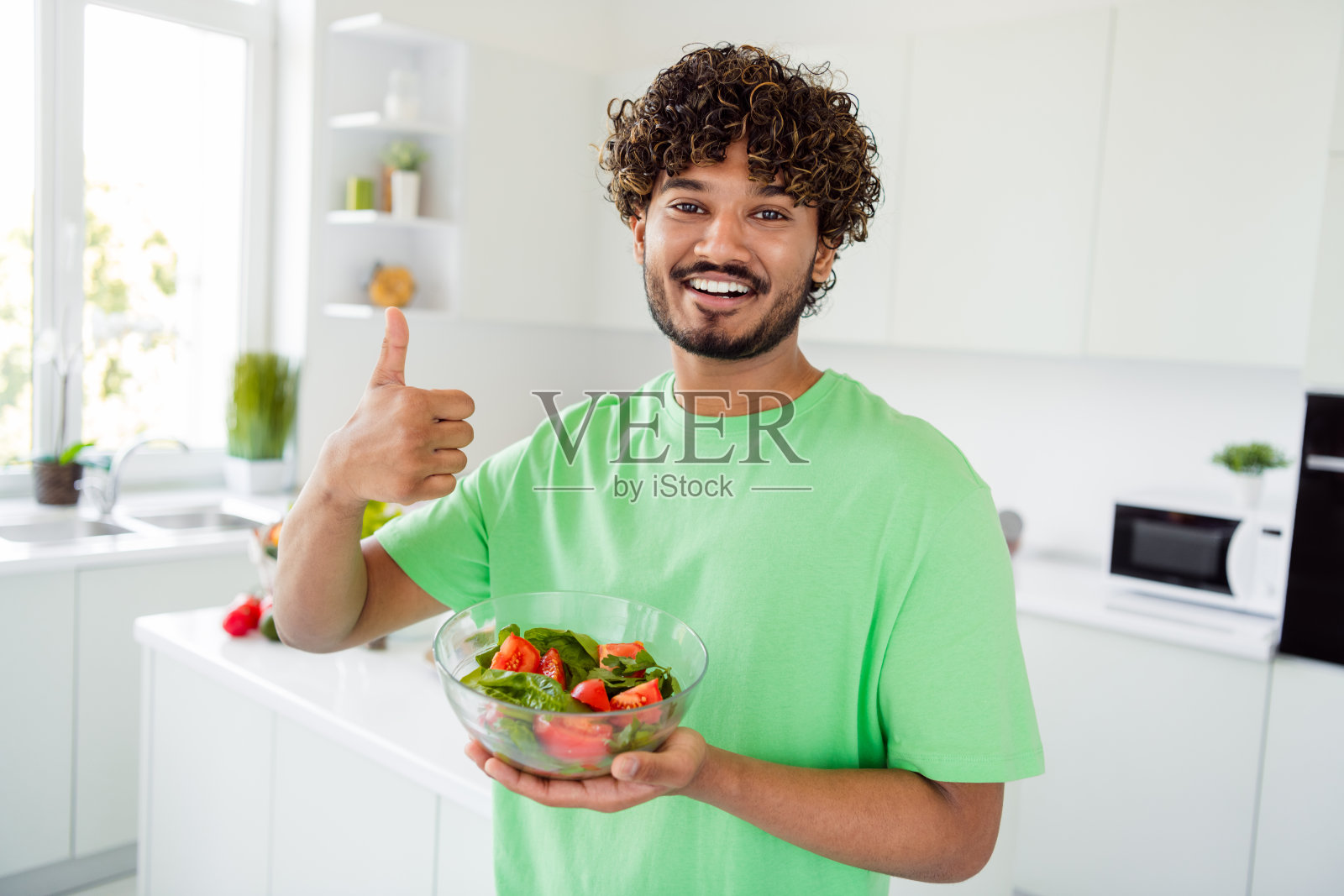 快乐的年轻男子在现代厨房里拿着一碗新鲜的沙拉，竖起大拇指，享受着明亮愉快的室内时光。照片摄影图片