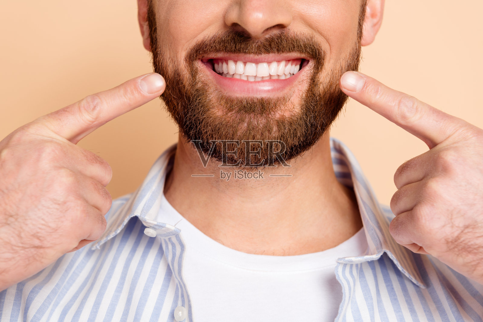 年轻男子自信地微笑，手势指向自己的牙齿，穿着休闲条纹衬衫，背景为米色。照片摄影图片