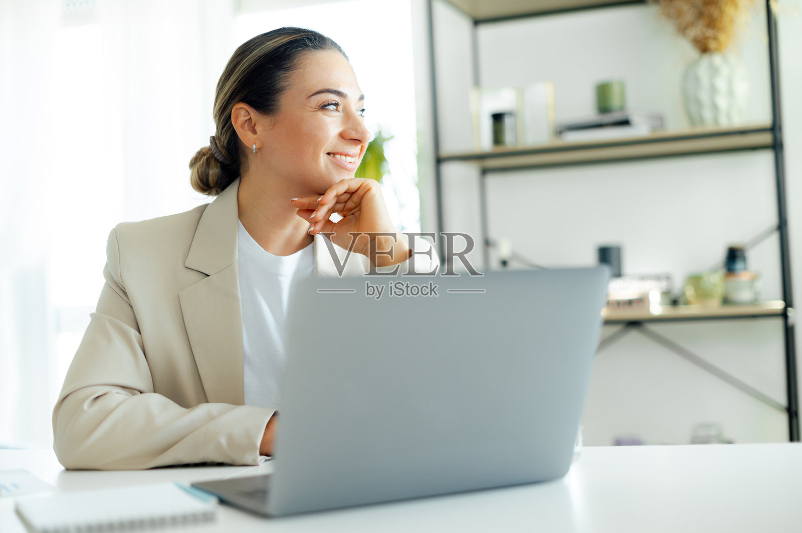 深思熟虑的积极美丽白人女商人，公司企业经理或首席执行官，坐在现代办公室的工作桌前使用笔记本电脑，微笑着望向屏幕之外。照片摄影图片