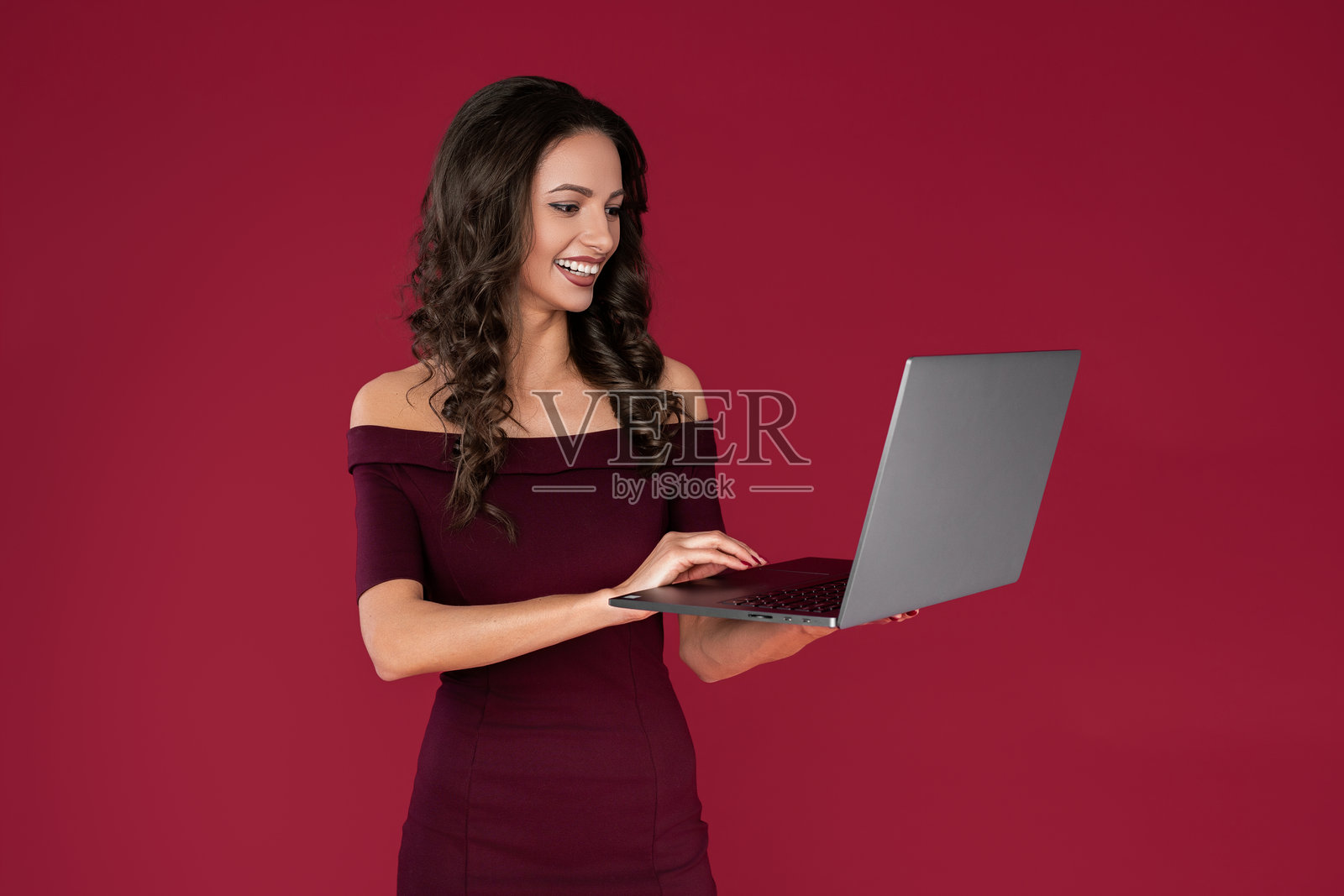 穿着深红色连衣裙的快乐优雅年轻女性在红色背景下用手指指着笔记本或电脑。照片摄影图片
