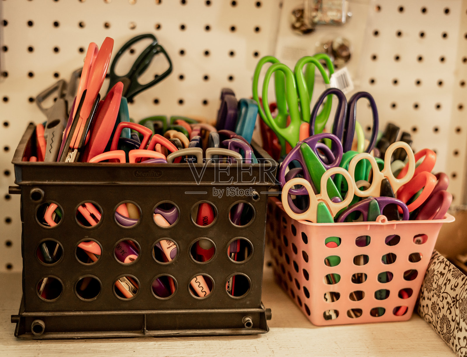 各种颜色鲜艳的手工艺和儿童剪刀存放在架子上的塑料篮子里，背景是挂板。这些剪刀形状各异，手柄明亮，通常用于艺术、手工艺或课堂环境。照片摄影图片